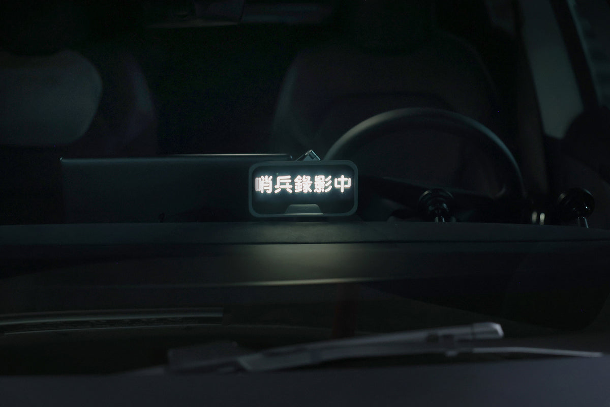 透過擋風玻璃,可見車內儀表板上放置著一款帶有發光中文字樣的 JOWUA 台灣萬用 MagSafe 磁吸燈。車內光線昏暗,背景中隱約可見方向盤的一部分。.