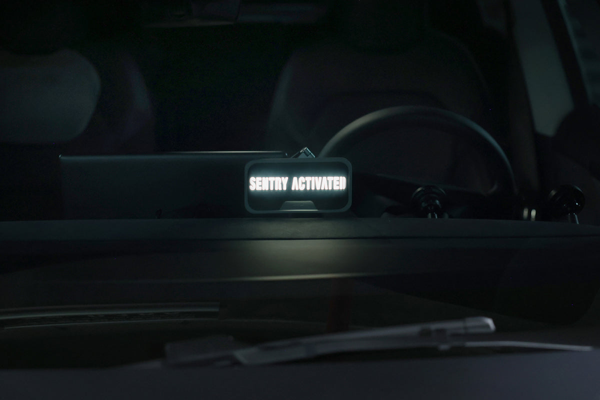 透過車前擋玻璃的近景畫面中,可見一個小型發光螢幕顯示著「SENTRY ACTIVATED」字樣,表示這輛停放中的車輛內部的安全系統已啟動。 在其旁,儀表板上可見一盞 JOWUA Taiwan 萬用 MagSafe 磁吸燈,它不僅與高科技車內裝完美融合,更提供多功能的磁吸照明,為駕駛人帶來額外的便利。.