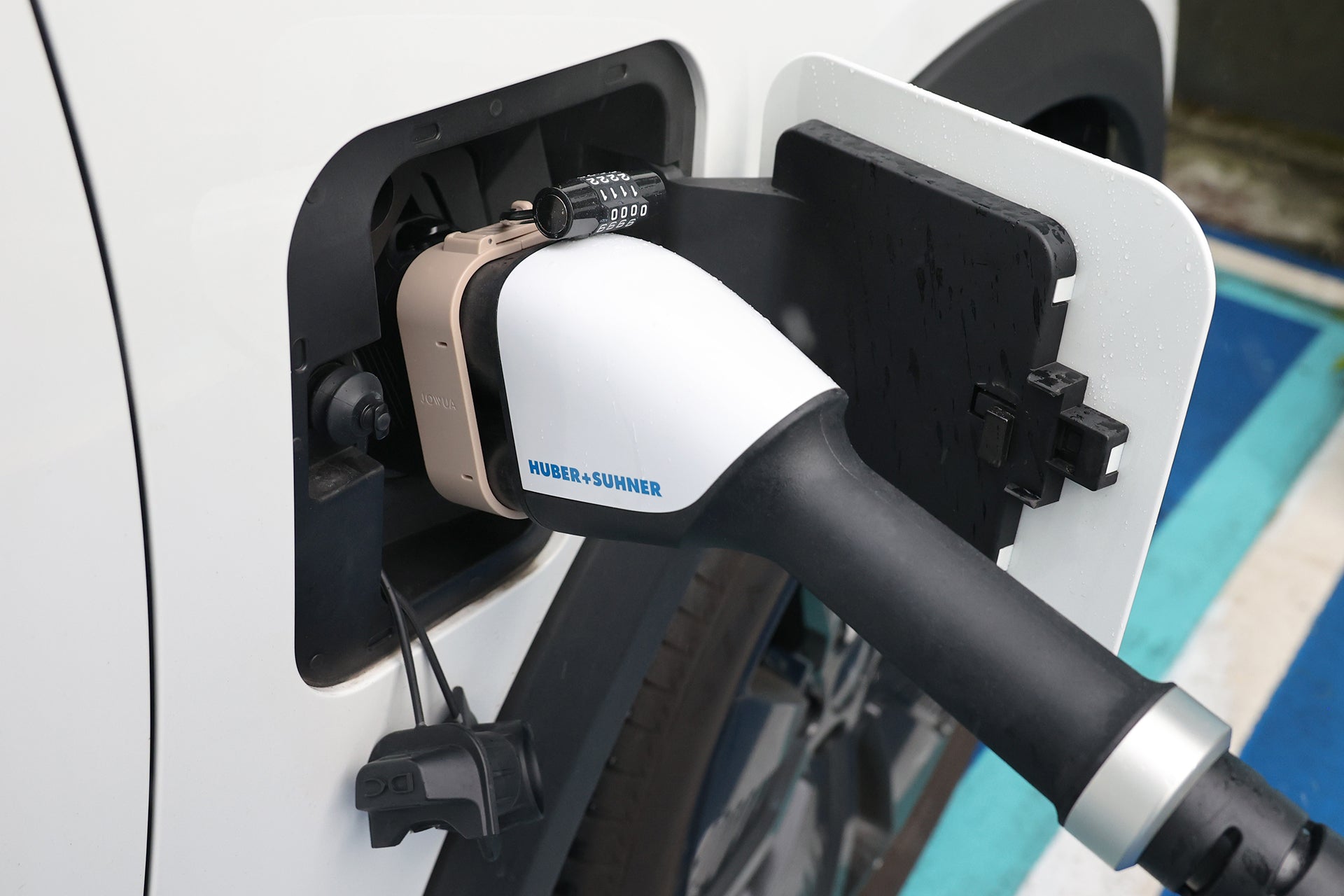 白色汽車充電口特寫,顯示 JOWUA Taiwan CCS2 to CCS1 充電防拔鎖扣已連接,並附有快速充轉接頭。背景為藍白線標示的停車區。.