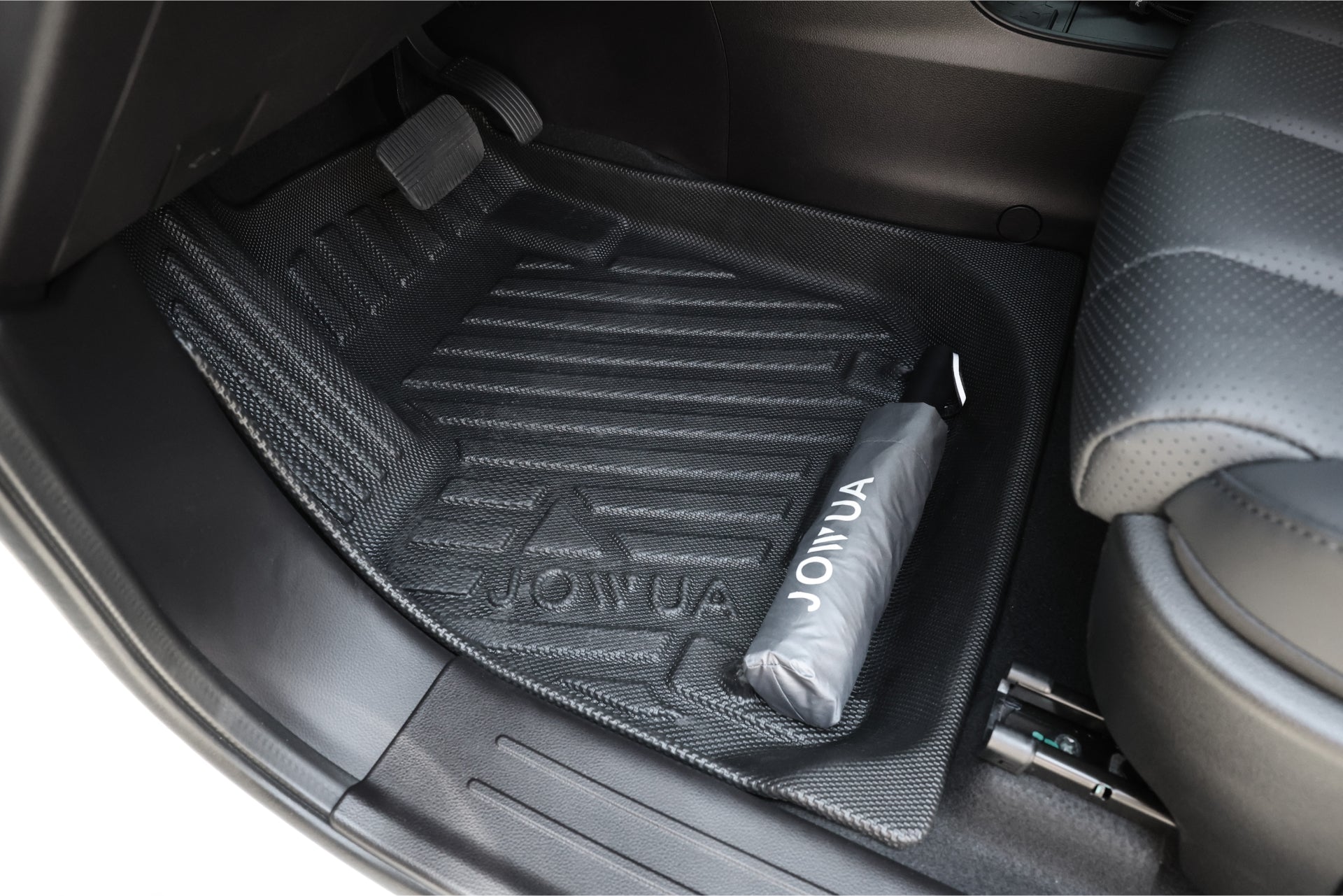 圖片展示了汽車內部,重點關注乘客側地板,其採用由 JOWUA Taiwan 生產的帶紋理的黑色「立體防水腳墊 (LUXGEN n⁷ 7人座)」。座椅旁邊有一把捲起的 Jowua 雨傘,體現了專利收納設計的最佳整理方式。.