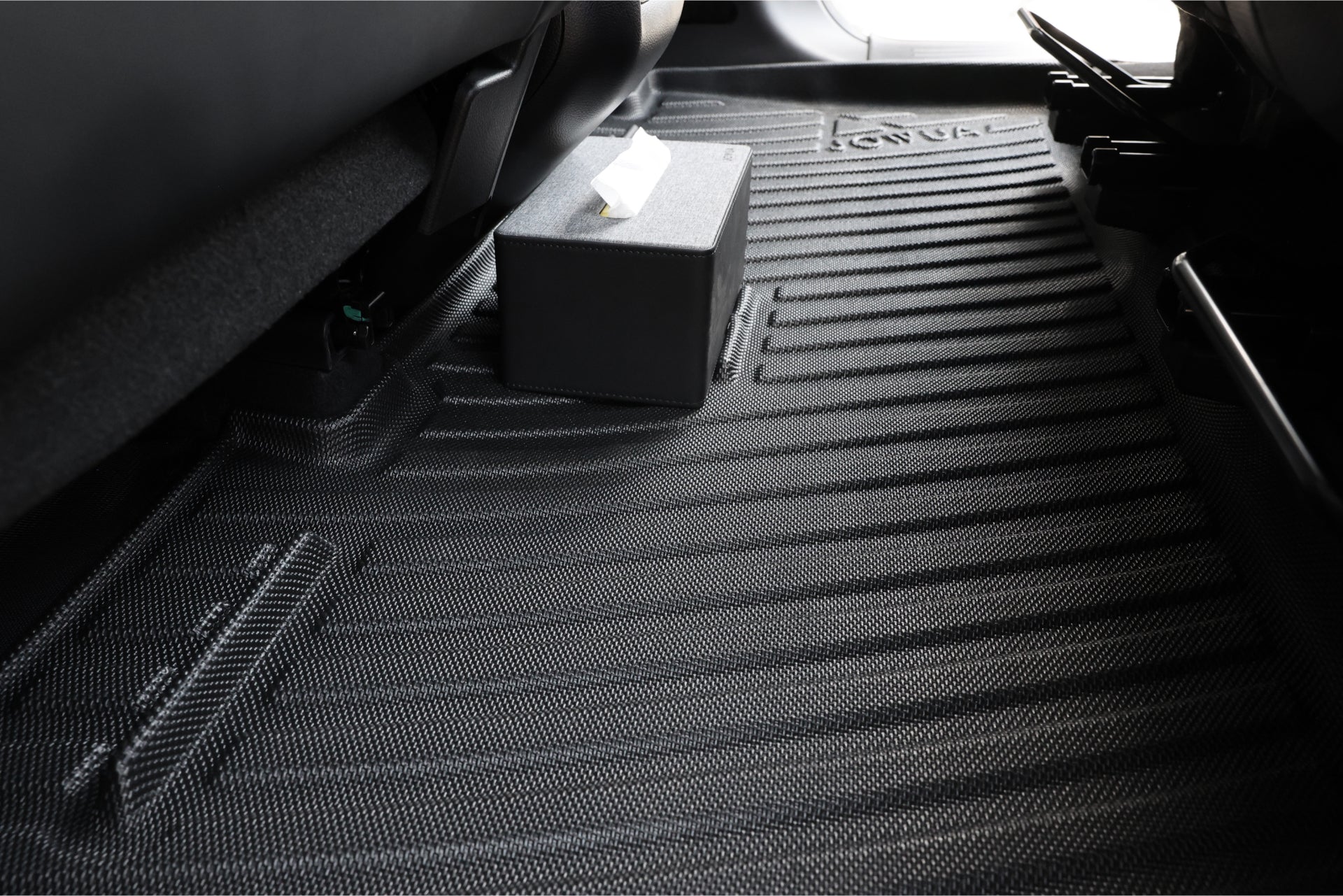 特寫 LUXGEN n⁷ 7 人座車的後座地板,配有帶紋理的黑色全天候 JOWUA 台灣墊。立體防水腳袊上放著一個裝有紙巾的小黑盒,其邊緣凸起,可防止灰塵和溢出。.