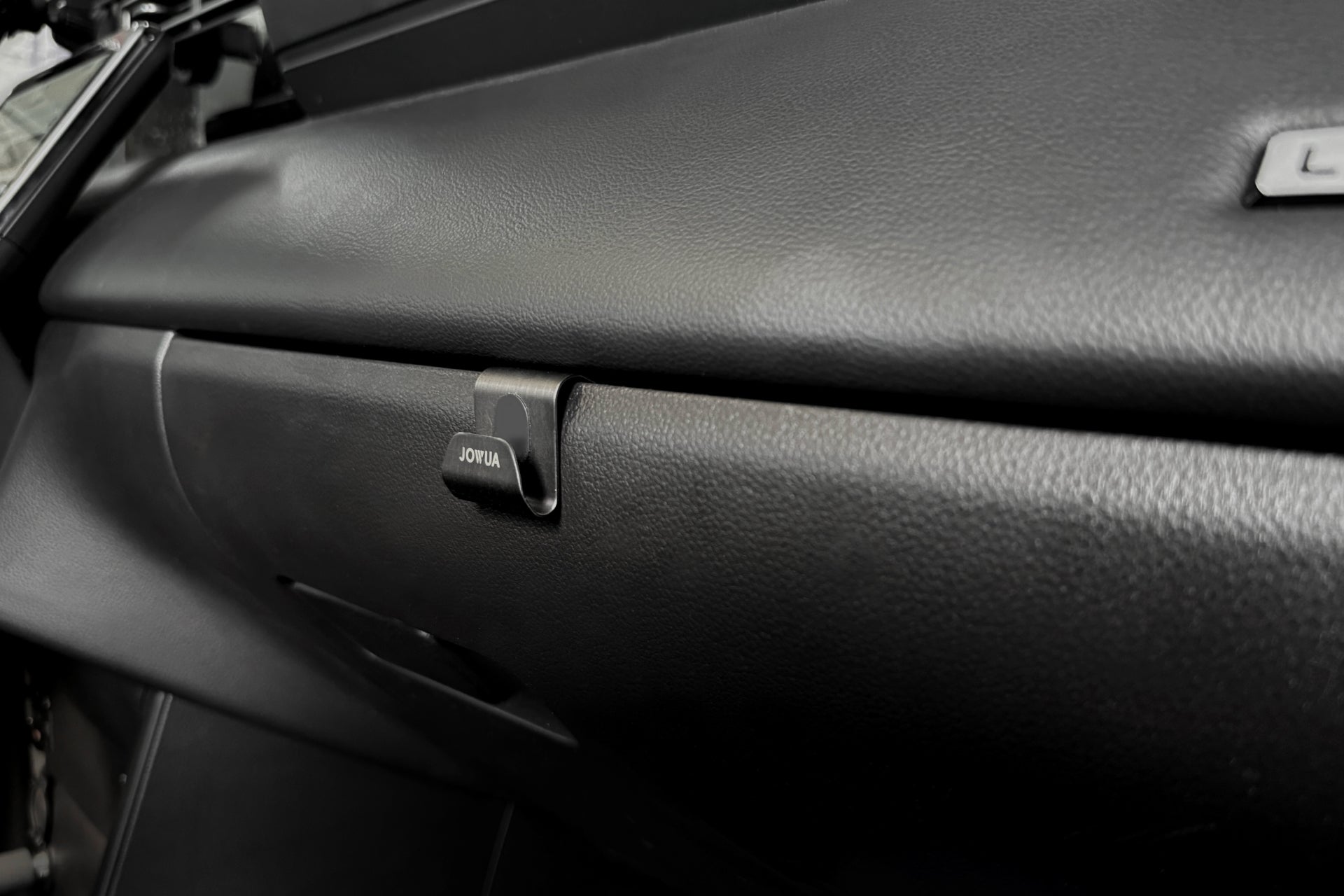 汽車黑色手套箱特寫,手柄上方貼有“JOWUA Taiwan”標籤。昏暗的內部突顯了帶有 LUXGEN n⁷ 手套箱掛鉤功能的紋理儀表板,可防止物品滑落。.