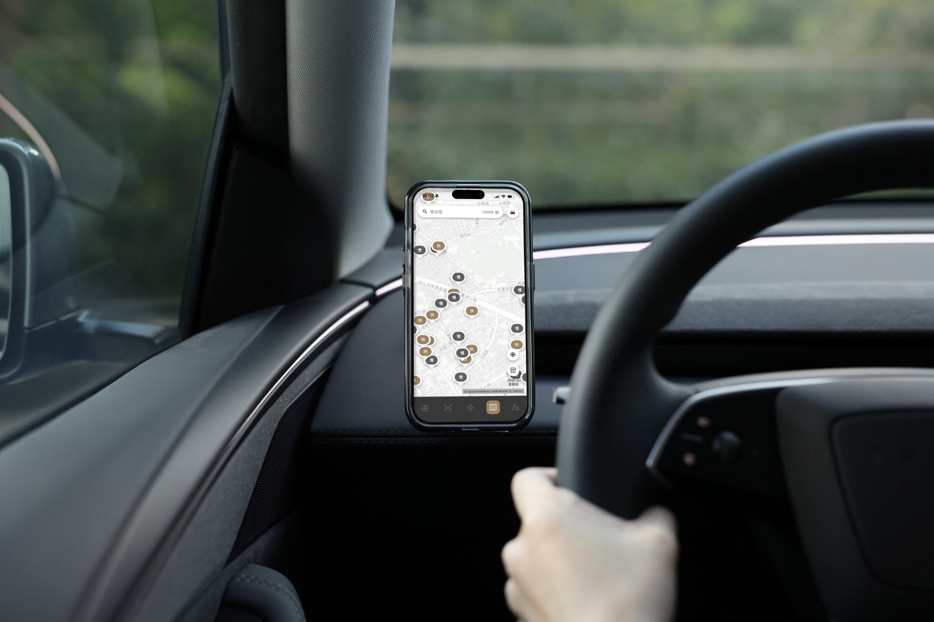 駕駛員單手握住方向盤,儀表板上安裝的 JOWUA Taiwan Qi2 製冷出風口 MagSafe 車架 (Model 3/Y 煥然一新版) 固定智慧型手機,顯示地圖上的插銷。車外可見模糊的綠意。.