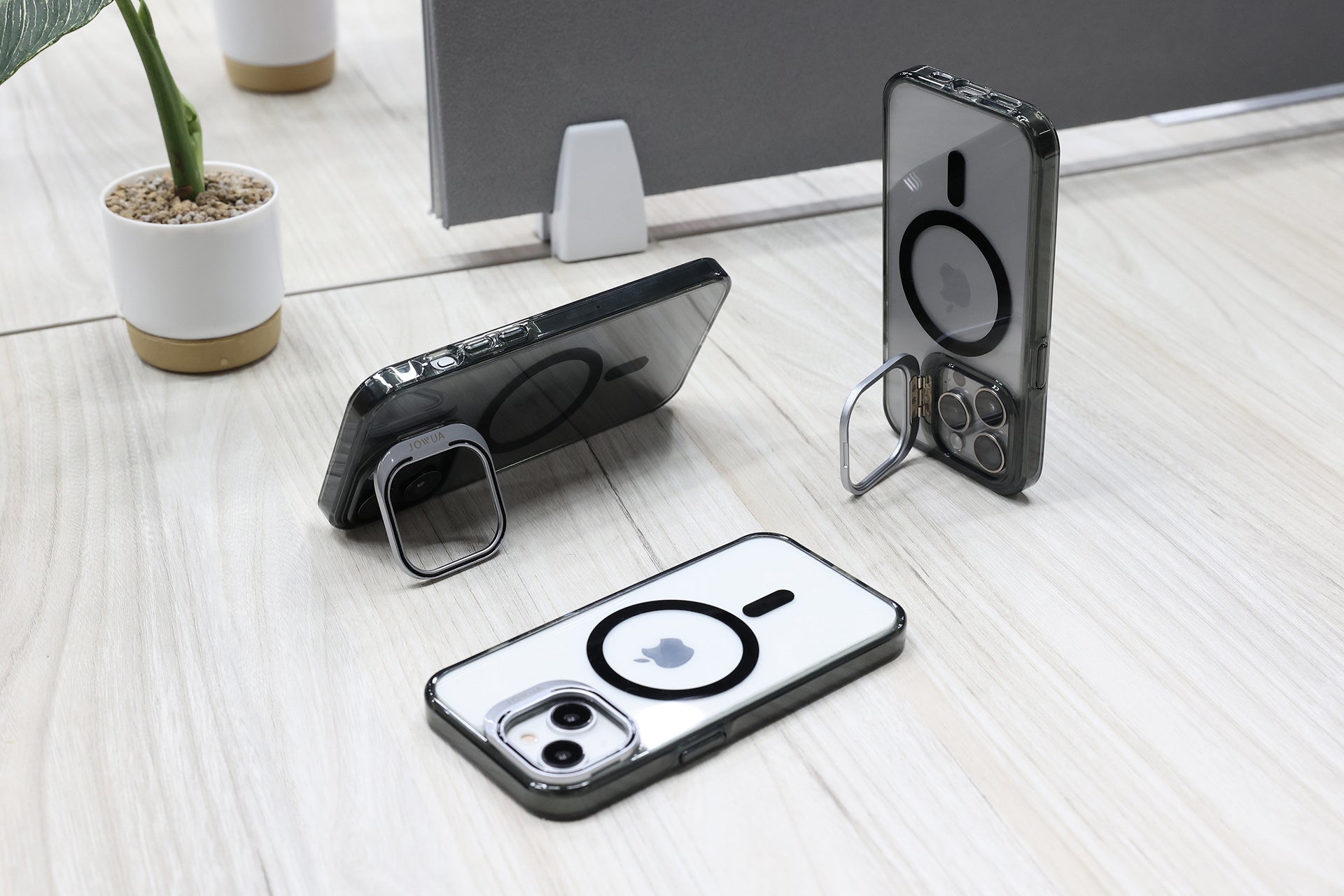 兩部裝有 JOWUA Taiwan MagSafe 透明支架保護殼 (iPhone 15/14/13 系列) 的 iPhone 放在一張輕木桌上,一張直立,一張平放,兩者都由金屬環支架支撐。小型盆栽點綴了整個場景。.