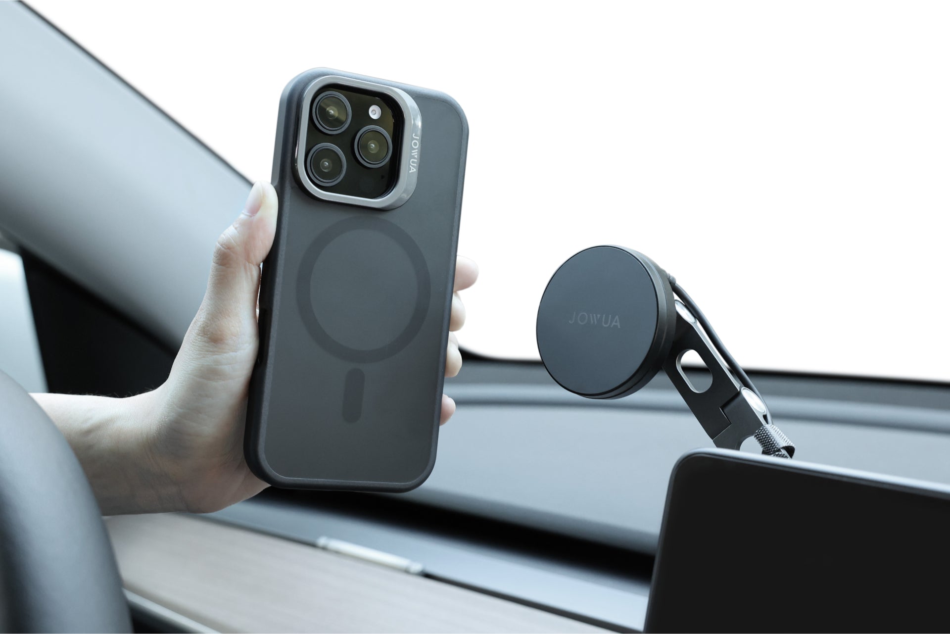 一手握著一部配有三個相機鏡頭的 iPhone 16,牢固地固定在車內 JOWUA Taiwan 的 MagSafe 透明支架保護殼+掛繩夾片組上。背景中的儀表板和擋風玻璃凸顯了技術與便利的無縫融合。.