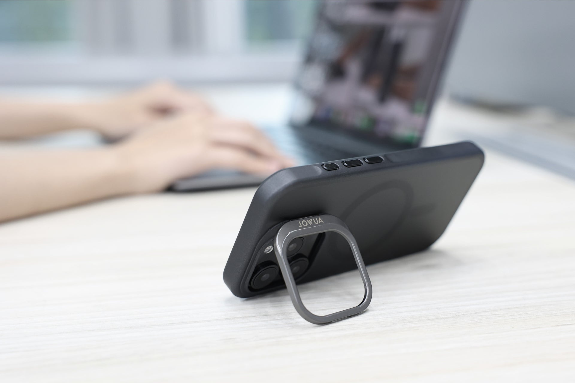 一台配有 JOWUA 台灣 MagSafe 透明支架保護殼+掛繩夾片組的 iPhone 16 支撐在桌上。背景中,一個人用模糊的雙手在筆記型電腦上打字。.