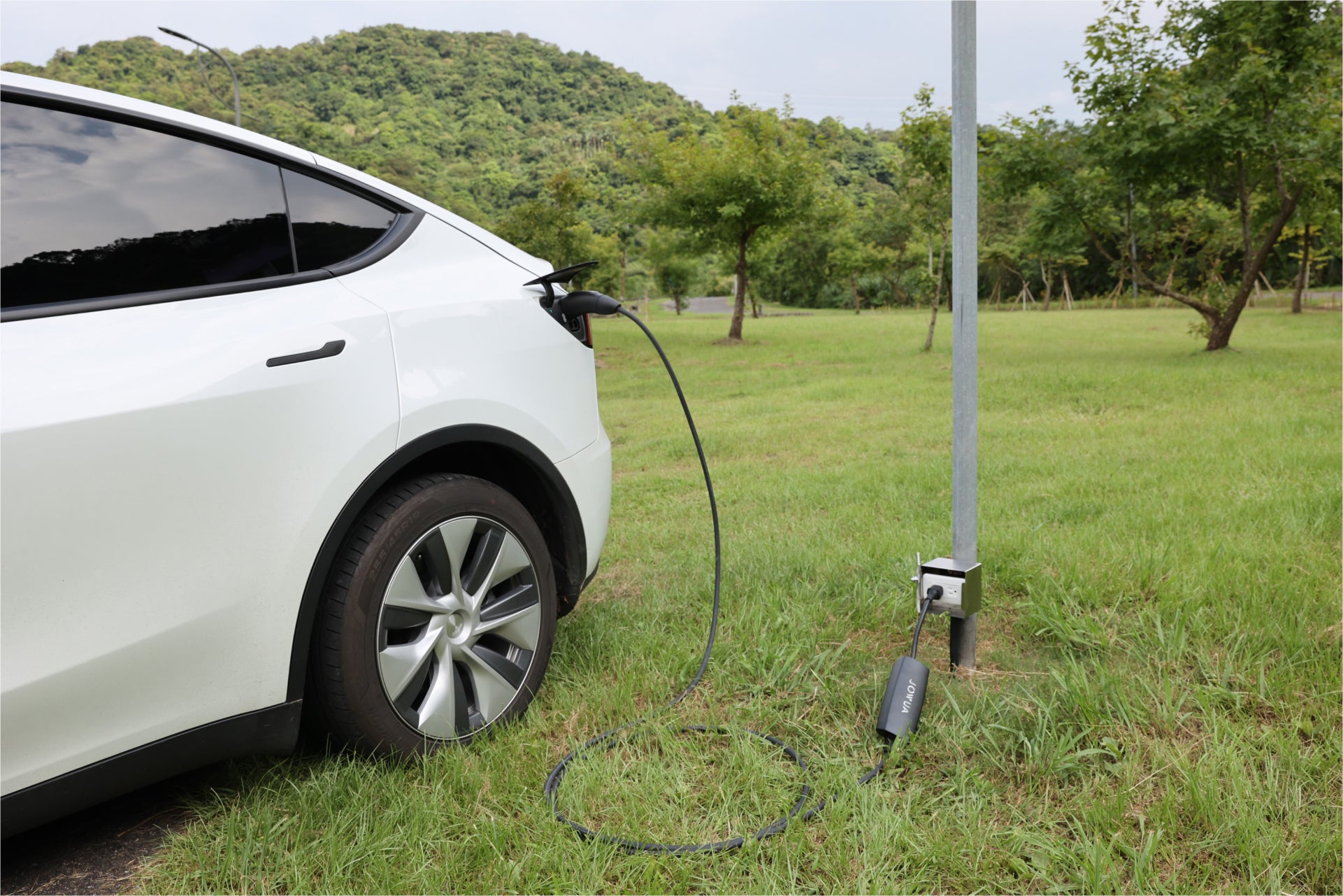 一輛白色電動車停在草地上,透過 JOWUA Taiwan 雙用行動充電器 (CCS2-AC Type-2) 7kW 充電,配備 NEMA 14-50/6-20 轉接器,安裝在金屬杆上,背景是綠樹和山丘。.