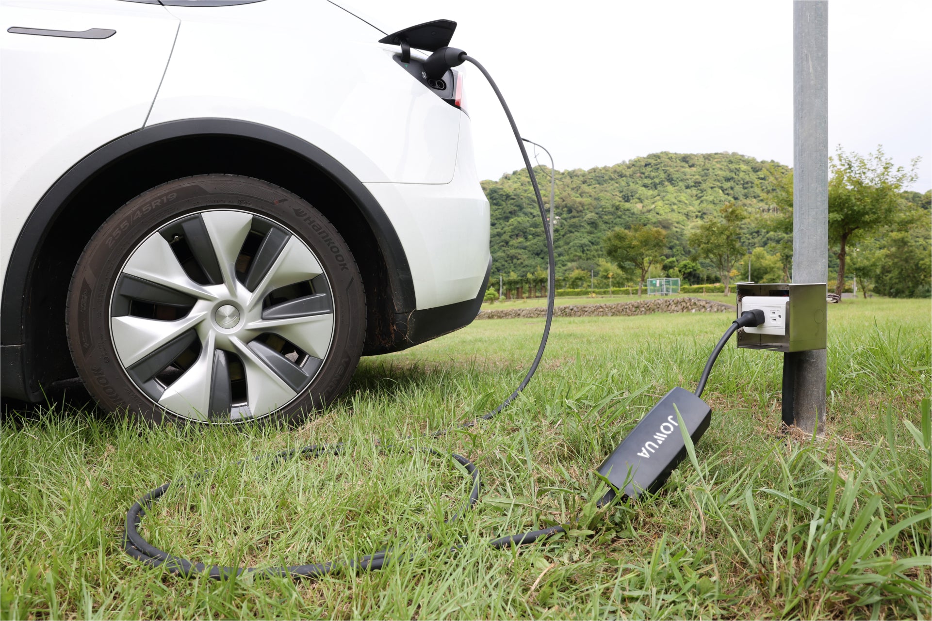 一輛白色電動車正在草地上充電,使用的是 JOWUA Taiwan 雙用行動充電器 (CCS2-AC Type-2) 7kW,配備 NEMA 14-50/6-20 轉接器,連接至金屬柱插座。背景為青山綠樹。.