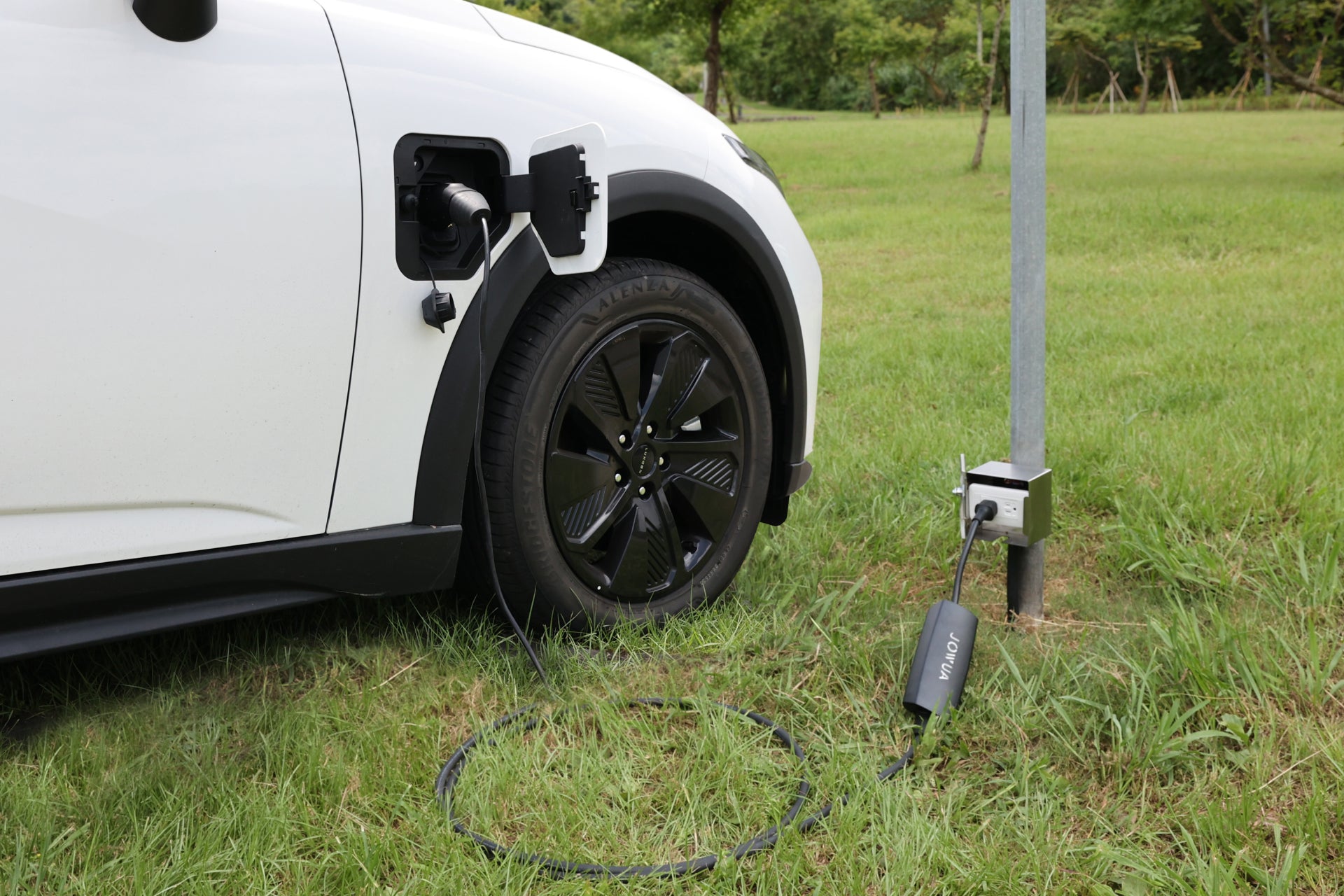 一輛白色電動車停在草地上,透過 JOWUA Taiwan 雙用行動充電器 (J1772/PHEV) 9.6kW 配備 NEMA 14-50/6-20轉接器組合進行充電,具有 IP55 防塵防水功能,可在戶外安全使用。連接線可插入左前輪上方的 J1772 充電座。.