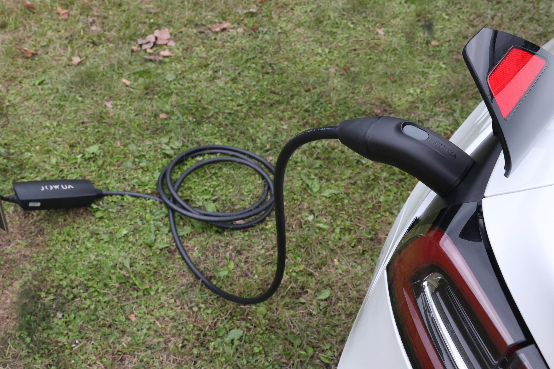一輛白色電動車在草地上使用 JOWUA Taiwan 雙用行動充電器 (TPC/NACS) 9.6kW|NEMA 14-50轉接器組合充電,具有 40A/9.6kW卓越充電效能;其黑色電纜在地上迴圈,連接至附近的充電器。.