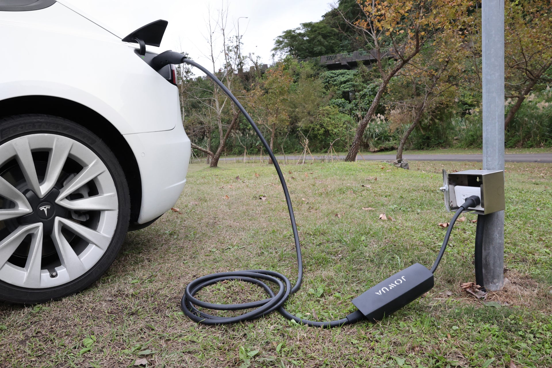 一輛白色 Tesla 停在草地上,使用 JOWUA Taiwan 雙用行動充電器 (TPC/NACS) 9.6kW|NEMA 14-50 轉接器組合充電。背景可見樹木與綠意。.