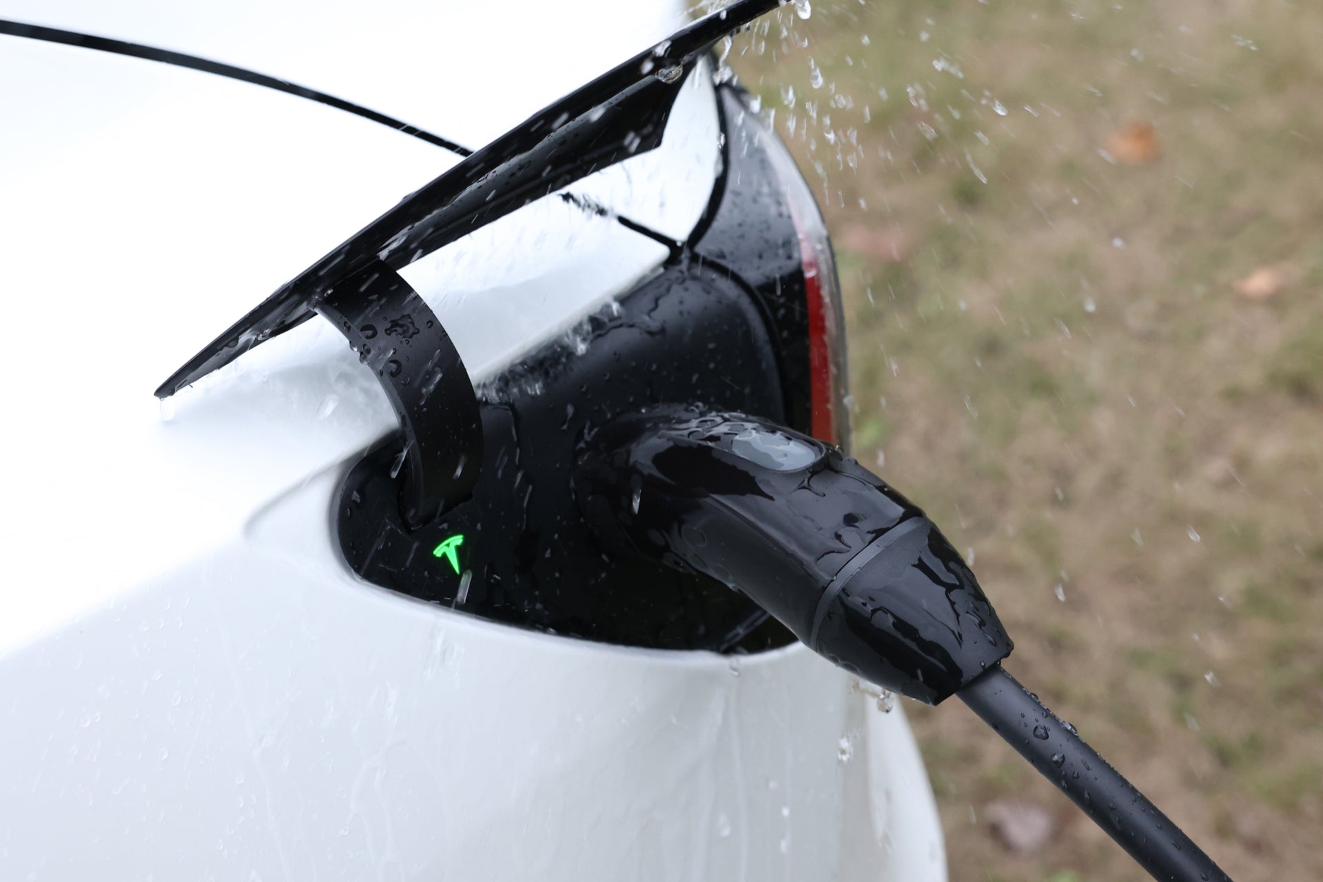 使用 JOWUA Taiwan 雙用行動充電器 (TPC/NACS) 9.6kW, NEMA 14-50轉接器組合在雨中為白色 EV 充電的戶外特寫,特寫特斯拉 TPC (NACS) 連接埠以及車輛和電纜上的水滴。.