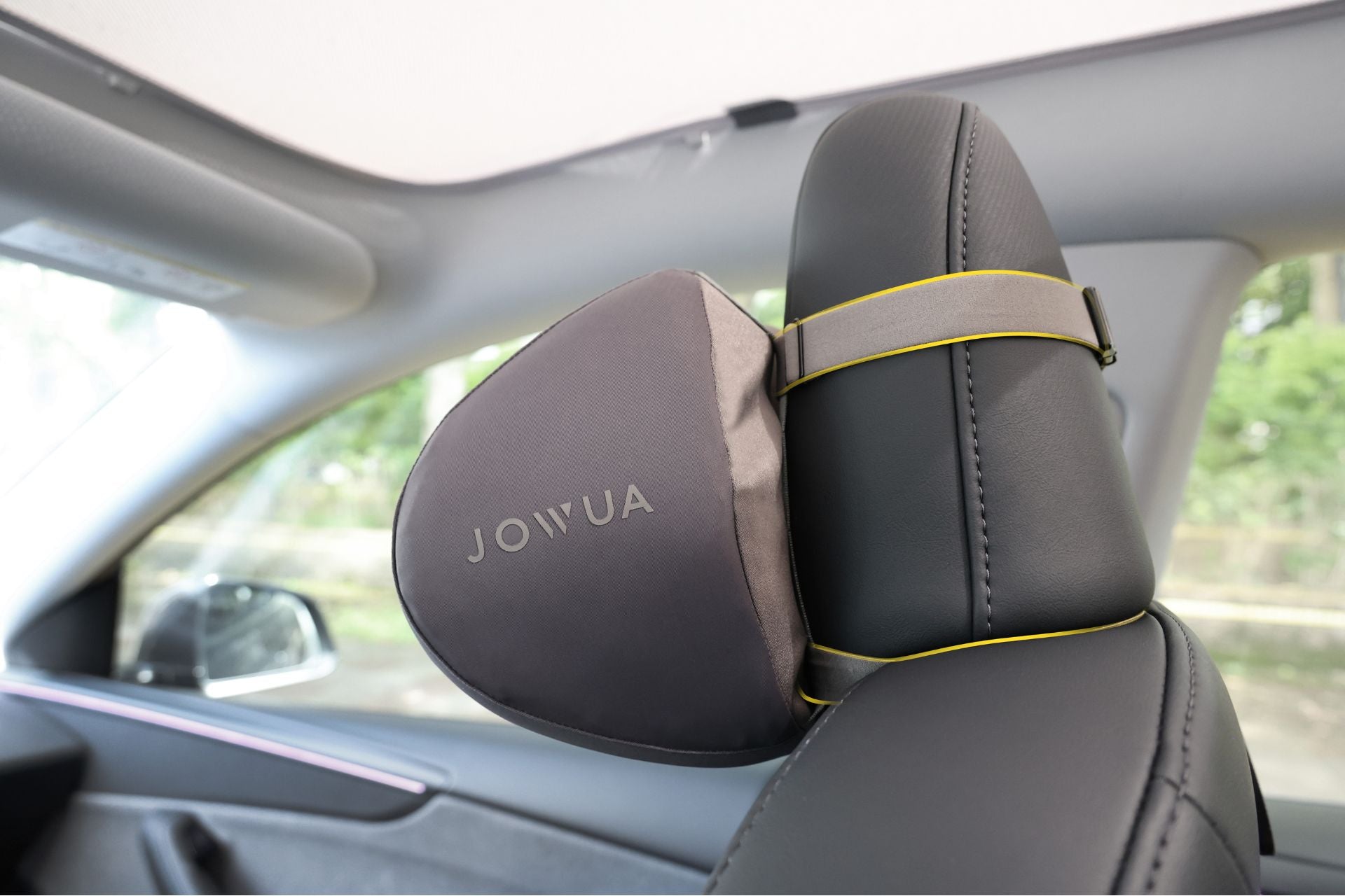 來自 JOWUA Taiwan 的灰色襯墊車用頭枕(兩入/一入)來自台灣 JOWUA 的灰色車用頭枕(兩入/一入),用黃色鬆緊帶固定在現代車輛的汽車座椅頭枕上,人體工學的設計,綠色的樹木從窗外可見。.