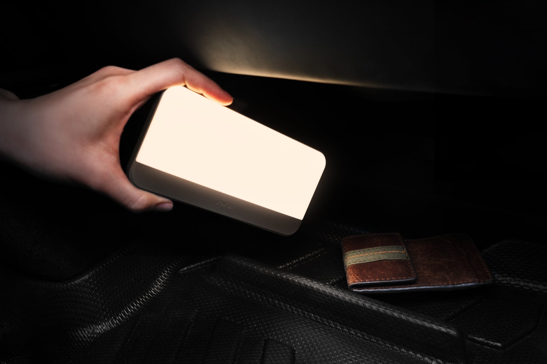 一隻手在黑暗中拿著明亮的 LED 燈,照亮了 JOWUA Taiwan 電墊專案【Model Y 煥新版|交車禮包】特斯拉 Model Y 專用的紋理墊子上的棕色錢包與綠色錶帶。.