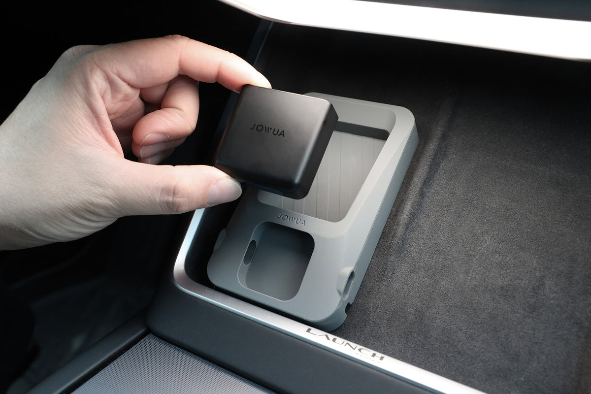 一隻手將一個標有「JOWUA 台灣 GPS 護盾」字樣、具備無線充電功能的小型黑色裝置,放入車內中控台區域的一個配套灰色托盤中。托盤與裝置完美契合。.
