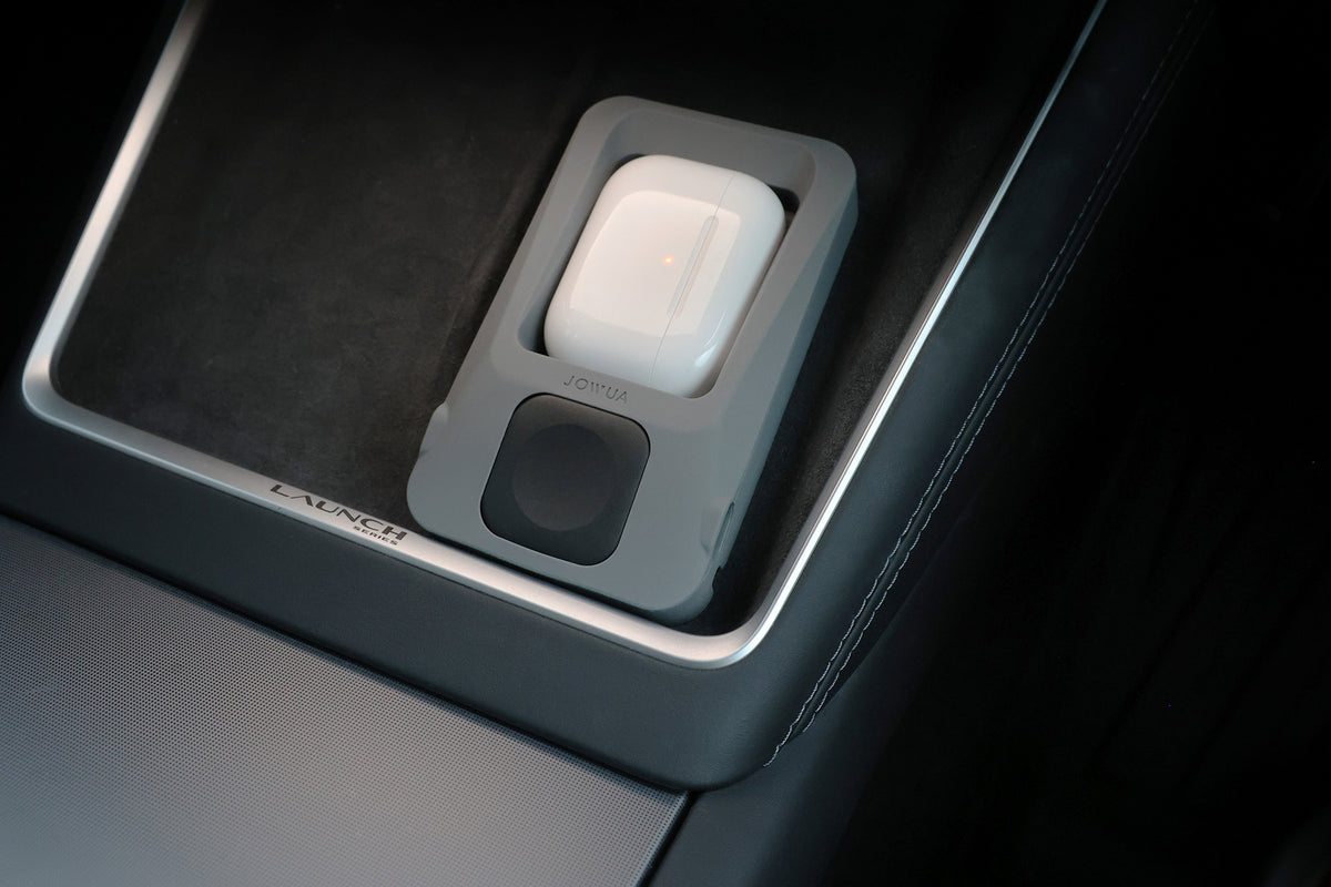 一個與 AirPods 無線充電相容的白色無線耳機充電盒,正置於車內中控台內的灰色 JOWUA 台灣製多功能無線充電底座+智慧手錶無線充電器上進行充電,附近標有「LAUNCH」字樣。.