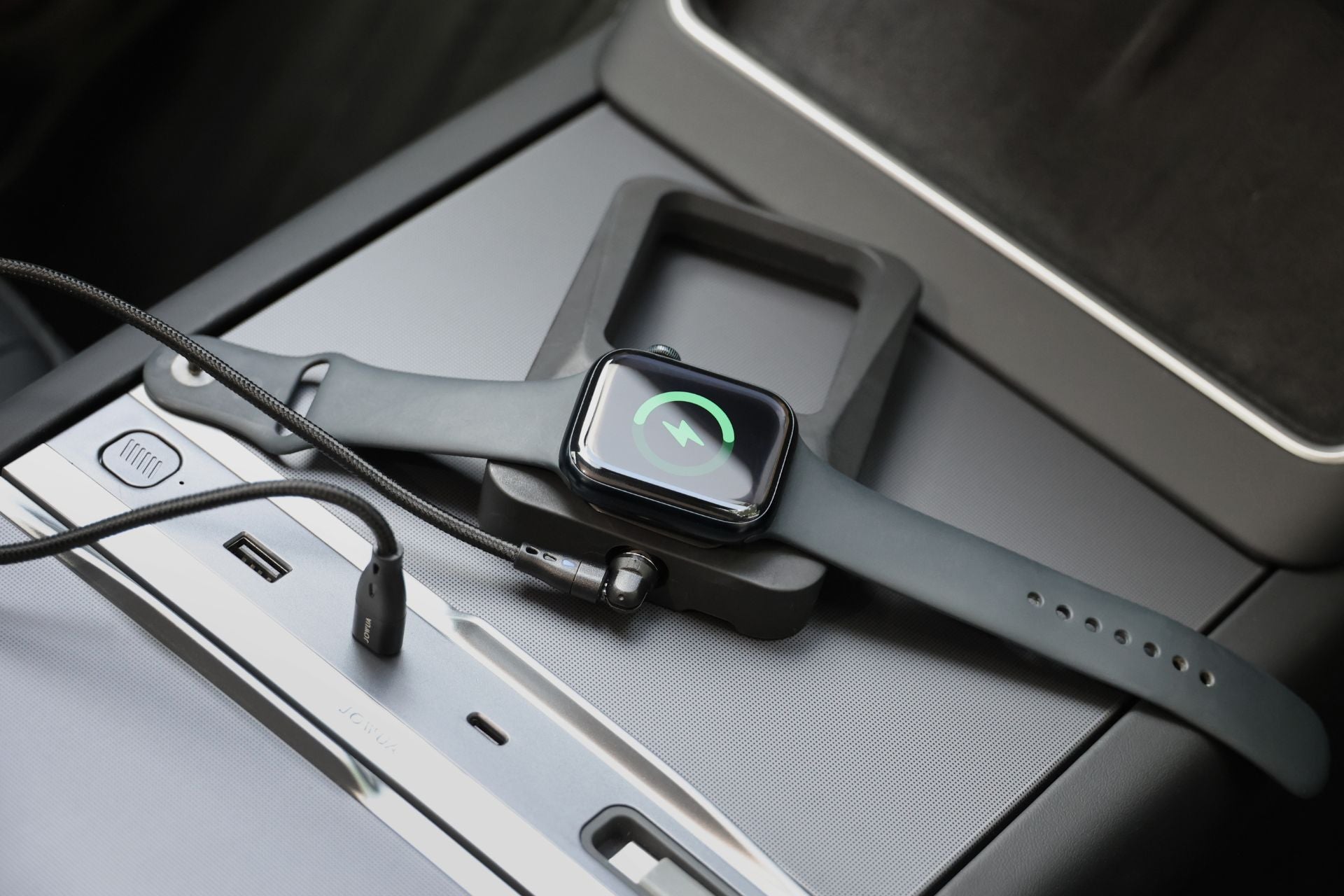 JOWUA Taiwan 多功能線無線充電底座+Apple Watch 無線充電器組合,黑色智慧型手錶正在汽車控制台上無線充電;附近有 USB 傳輸線和充電埠,手錶螢幕顯示充電符號。.