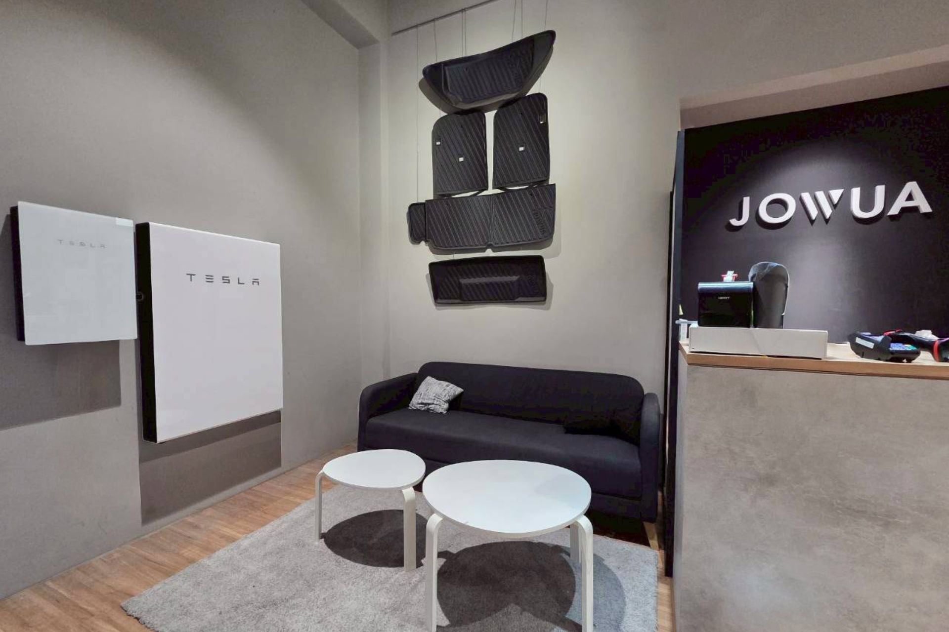 現場展示 JOWUA Taiwan Powerwall、黑色沙發、兩張白色圓桌、遮陽簾,以及「JOWUA」接待桌。牆上展示的智慧能源。所有控制都可透過 Tesla APP 輕鬆進行。.
