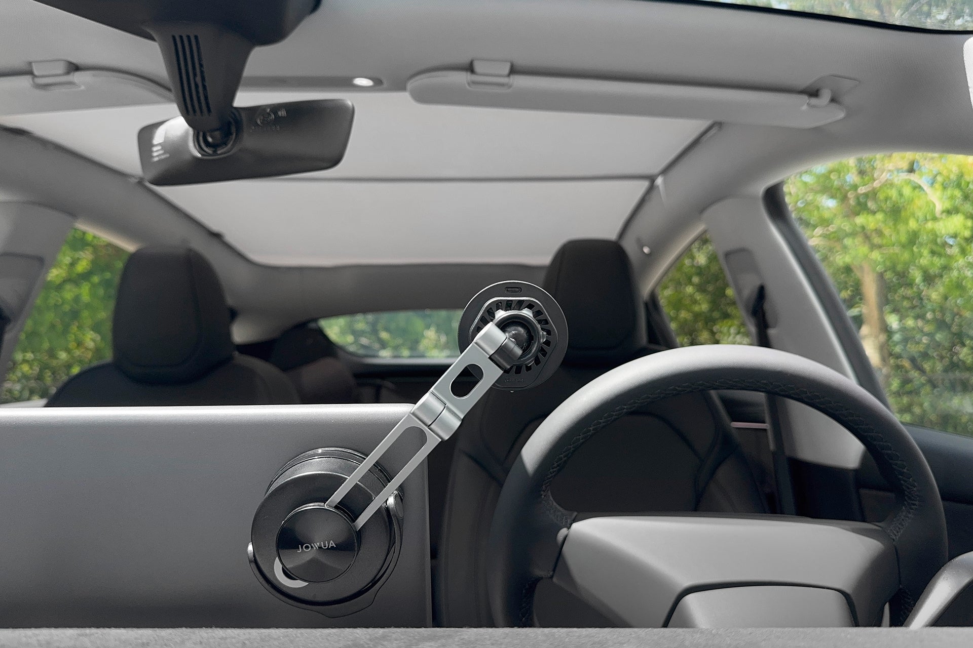 汽車內部特寫,顯示 JOWUA Taiwan Qi2 製冷六維隱藏車架 (MagSafe 款) 連接到擋風玻璃上,兩個前座、方向盤和後視鏡清晰可見。透過車窗可看到外面的樹木。.