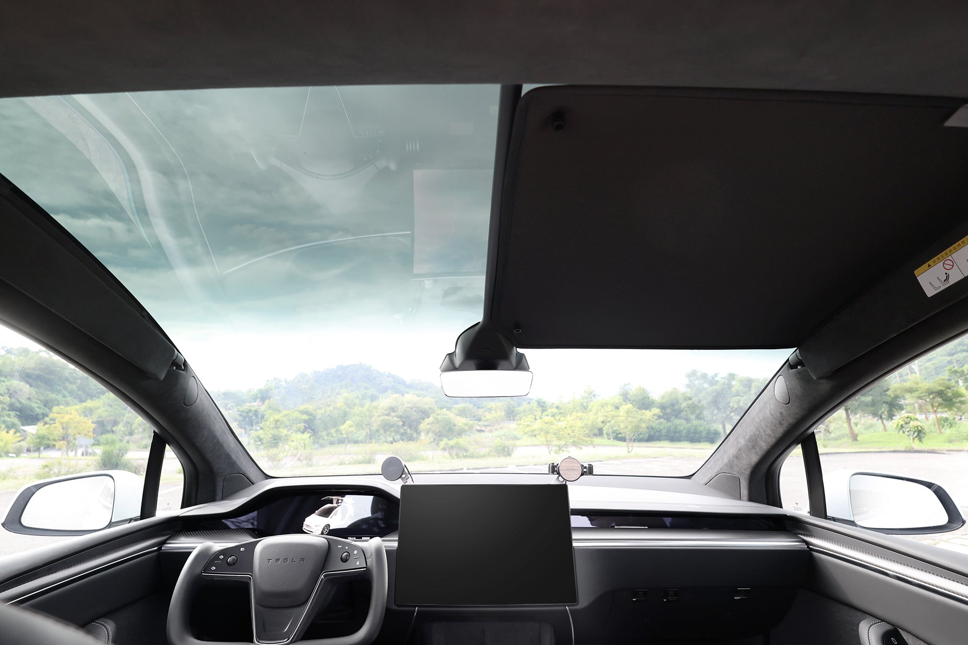 從配備全景擋風玻璃、觸控螢幕和側後視鏡的現代汽車上看出去的景色。外面是樹木和雲。JOWUA Taiwan 分離式遮陽簾 (Model X) 可防紫外線,非常適合具有隔熱車頂或鷹翼門專用功能的汽車。.