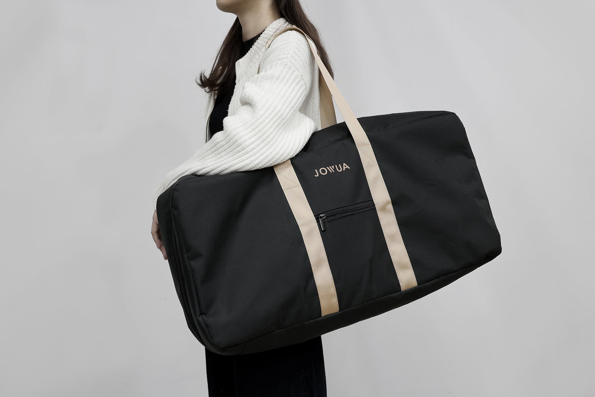 A person in a white sweater holds a JOWUA Taiwan 前車廂 & 滑板車專用收納袋-33L大容量,黑色配米色手把及前拉鍊袋,適合特斯拉前車廂收納與攜帶JOWUA折疊電動滑板車。背景為淺色純色。.