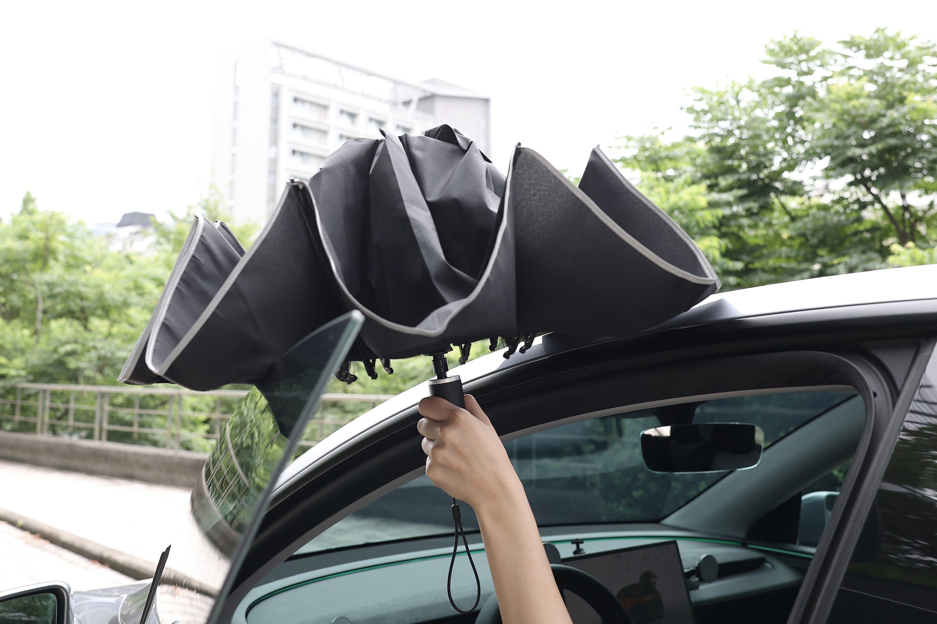 一個人拿著半封閉的黑色 JOWUA Taiwan 一鍵開收電動傘在汽車窗外,背景是綠意和建築物。.
