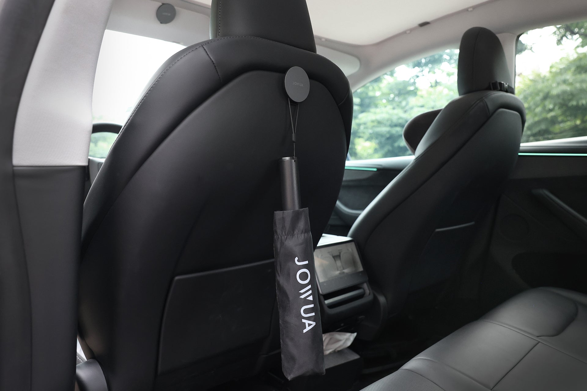 在現代車輛中,黑色的 JOWUA Taiwan 一鍵開收電動傘懸掛在黑色真皮汽車座椅後方,從車窗可以看到樹木和綠地。.