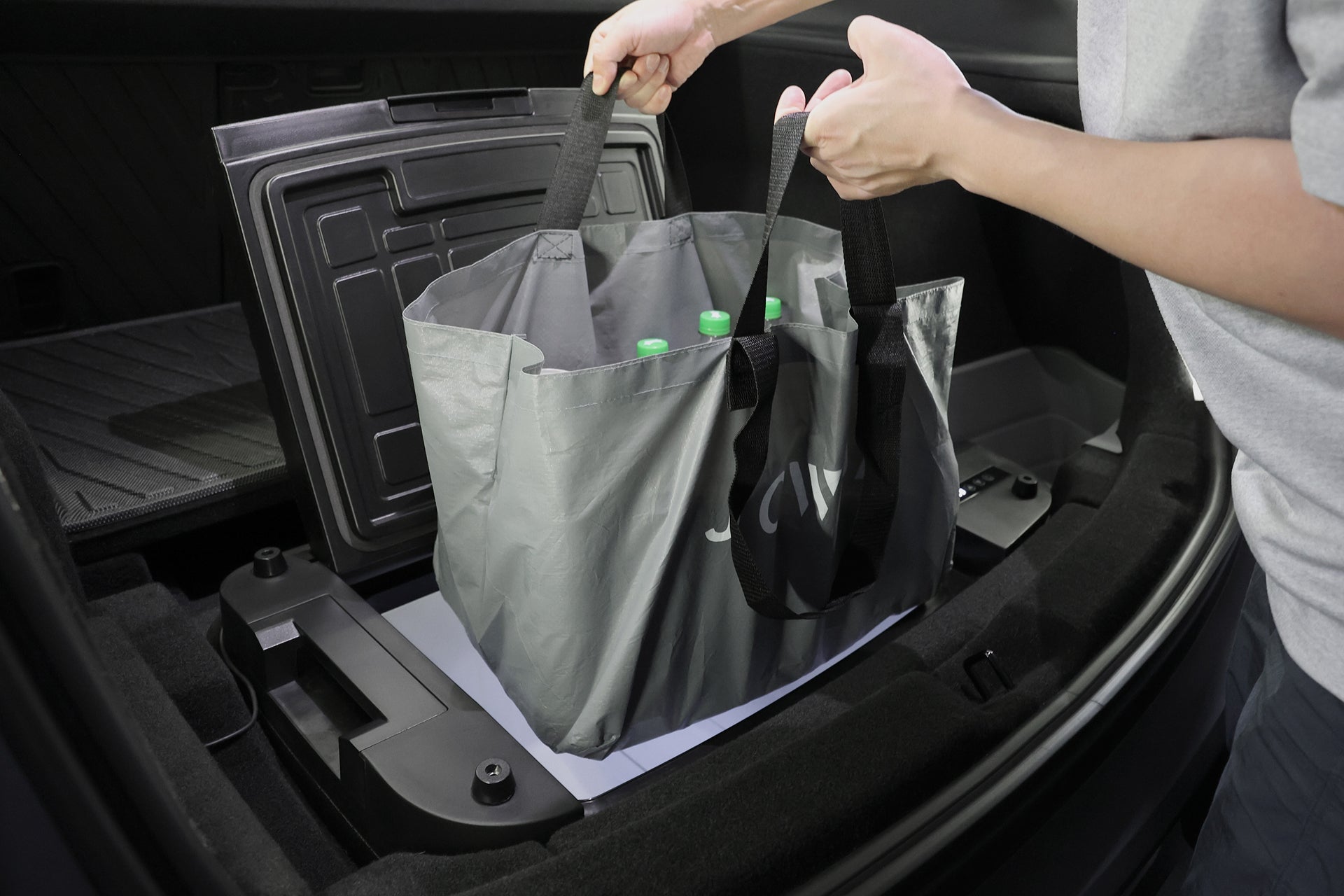 一個人將一個裝有水瓶的灰色可重複使用購物袋放進汽車後備箱,旁邊是一個 JOWUA Taiwan Type-C 車用行動冰箱 (Model Y全車系),方便隨身冷卻。.