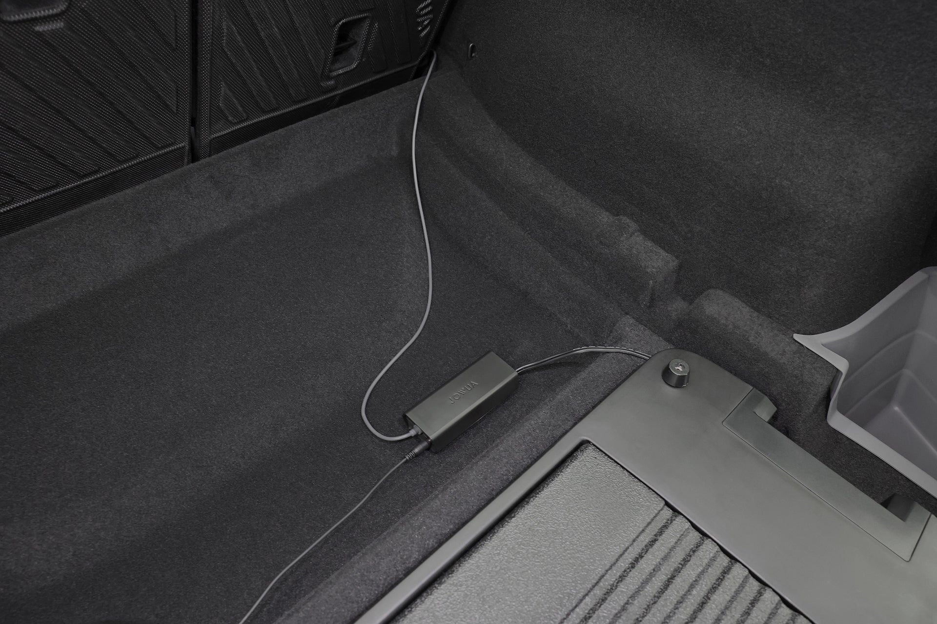 汽車後備箱的特寫照片顯示,JOWUA Taiwan 的 Type-C 車用行動冰箱 (Model Y 全車系) 擺在地毯上,與充電線連接,旁邊還有部分塑膠儲物箱和可摺疊座椅。.