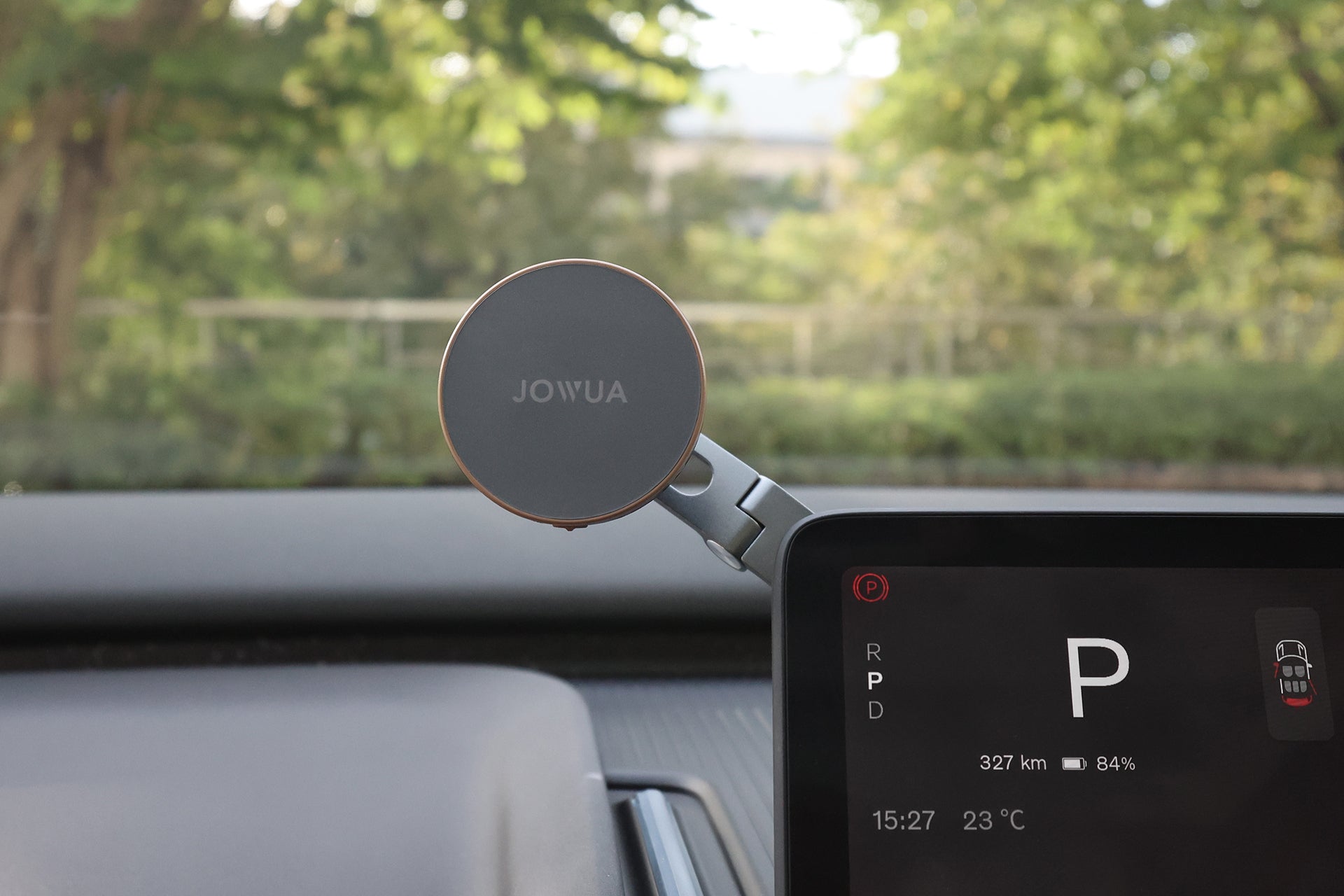 在汽車儀表板上安裝了 JOWUA Taiwan Qi2 製冷六維隱藏車架 (MagSafe款)。顯示器顯示停車模式、327 公里續航里程、84% 電量。透過擋風玻璃可以看到綠樹。.