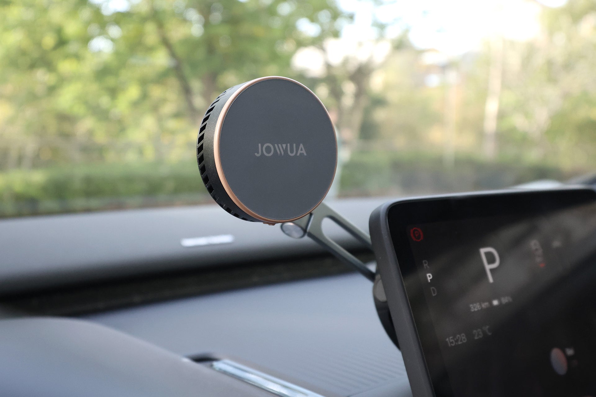 一個圓形的 JOWUA Taiwan Qi2 製冷六維隱藏車架 (MagSafe 款) 裝在汽車儀表板上,靠近數位顯示器,背景是透過擋風玻璃看到的綠色模糊戶外景象。.