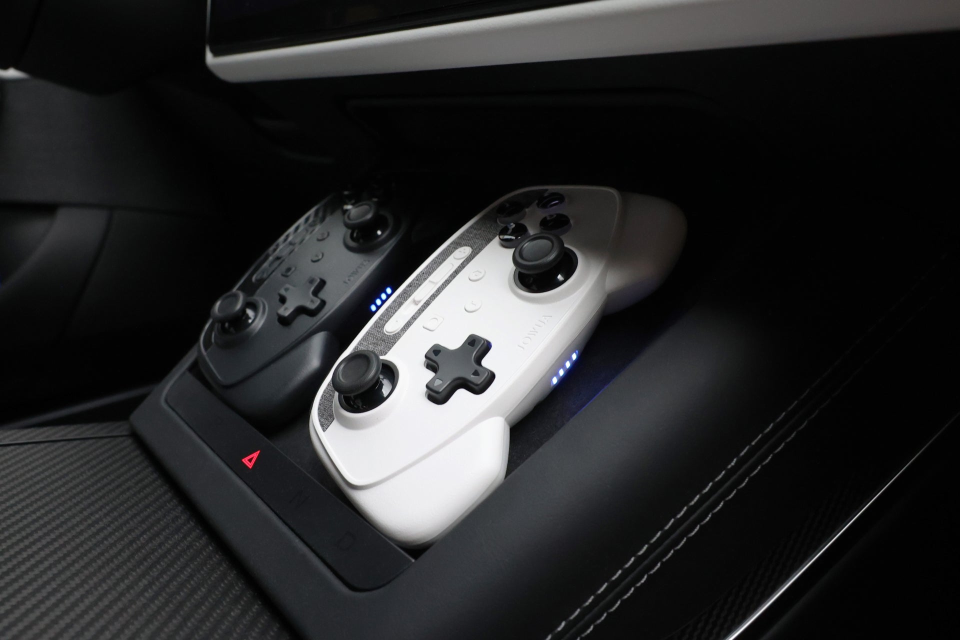 兩支 JOWUA Taiwan 無線充電雙搖桿組合 (Model 3/Y) 遊戲控制器,一黑一白,並排放置在汽車的碳纖維風格中控台上,展示它們與特斯拉手套箱集線器的相容性。.