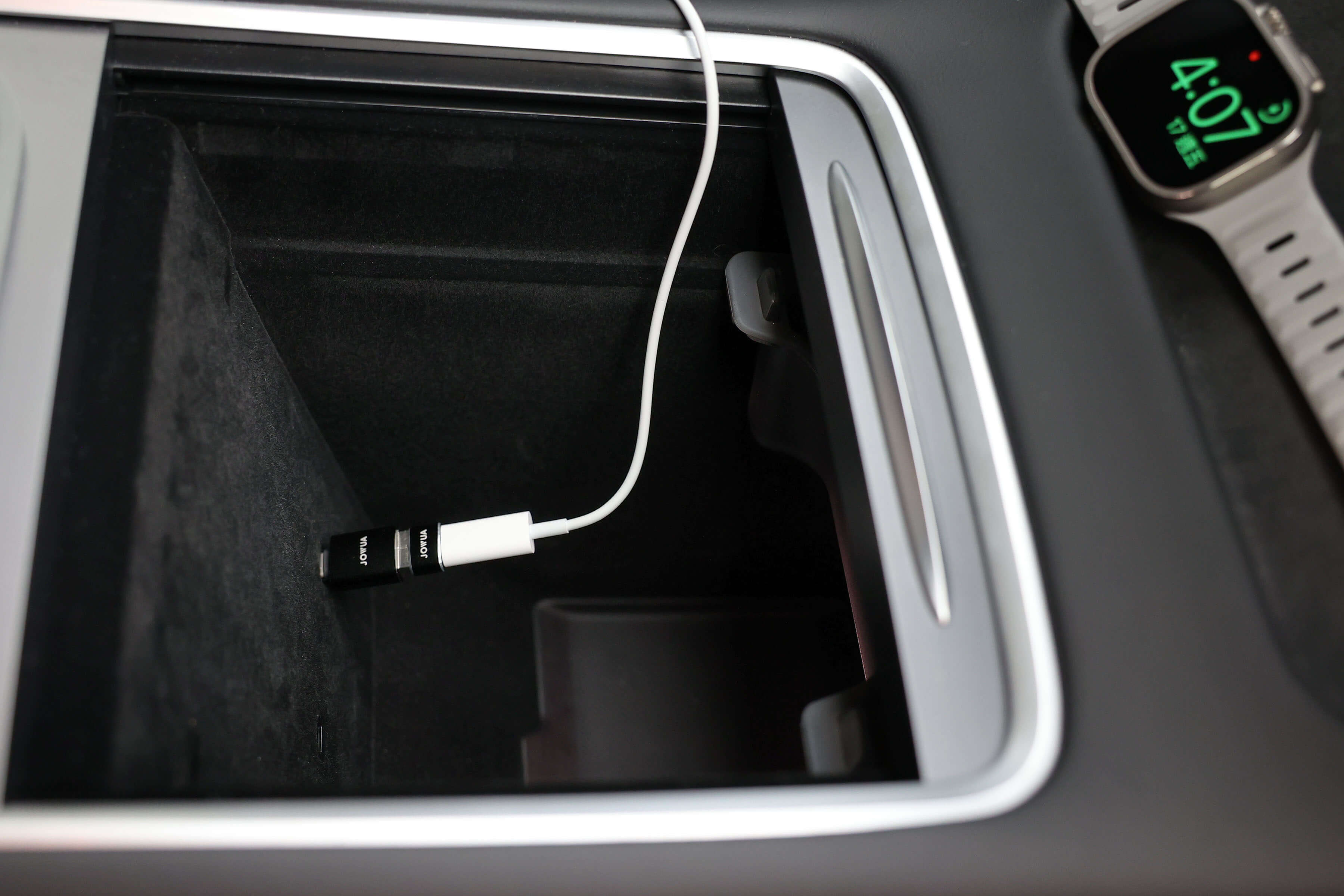 在汽車的內部儲物箱中,一個白色的 USB-C 轉接器連接到 USB 連接埠。扶手附近,有一款由 JOWUA Taiwan 出品的 Apple Watch 充電套件,其中裝有一塊 Apple Watch,其灰色錶帶上顯示“4:07”,凸顯了其時尚的設計和耐用的材質。.