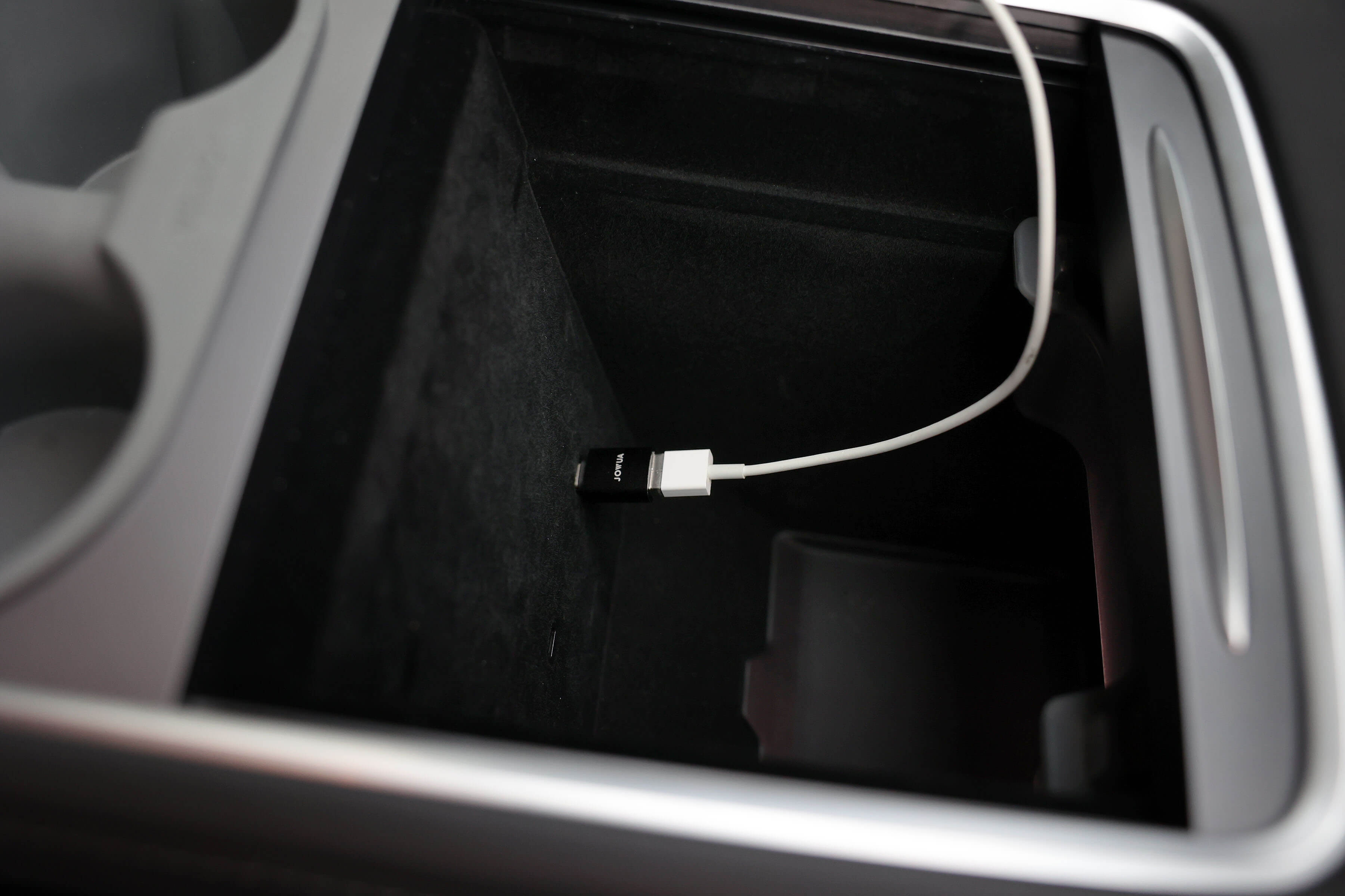 JOWUA 台灣 Apple Watch 充電套件配備耐用的 USB-C 轉接器和白色電纜,可插入汽車中控台杯架旁的隔間。內部很暗,電纜部分捲在邊緣。.