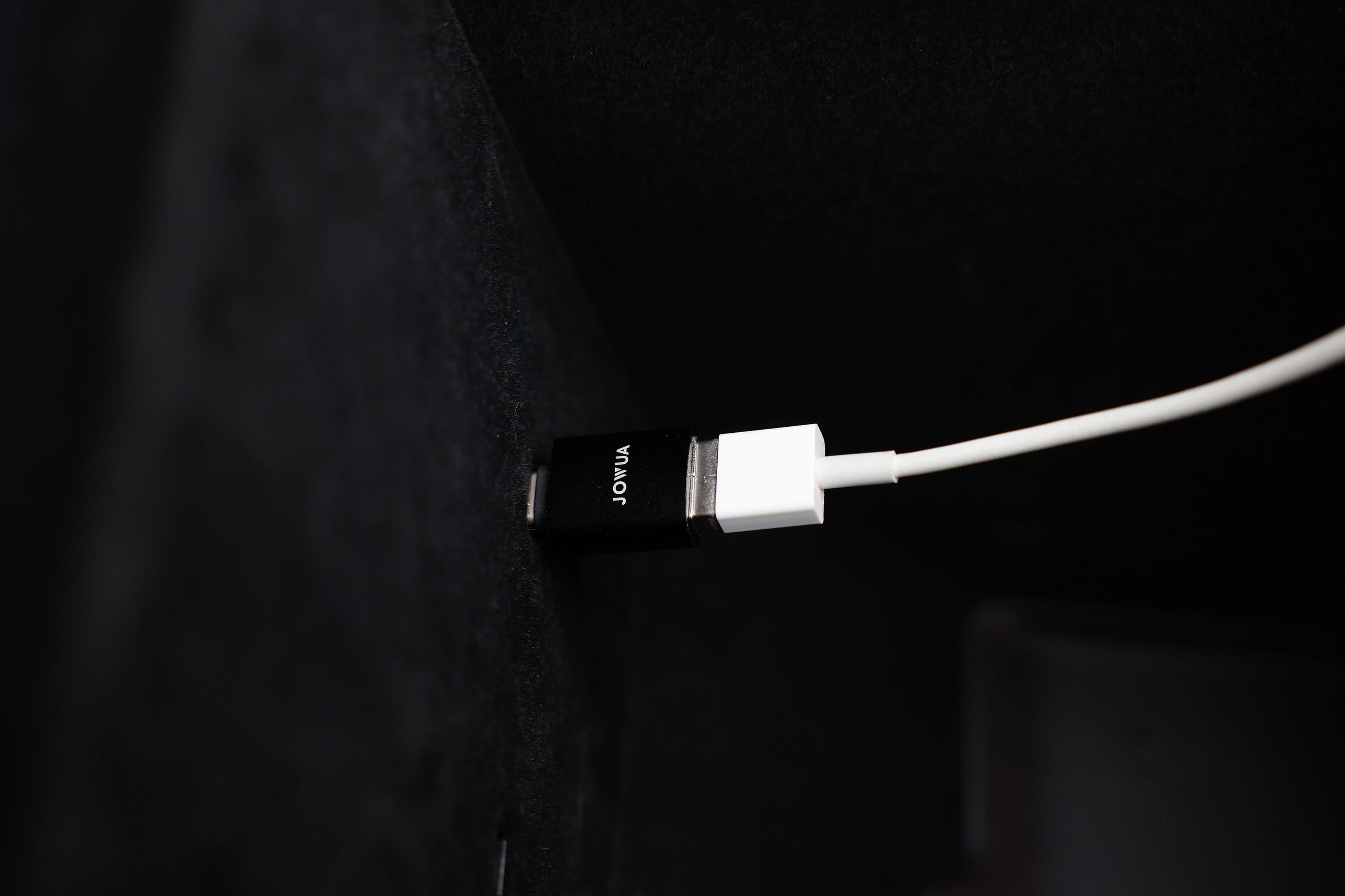 特寫:白色 USB 線連接到 JOWUA 台灣 Apple Watch 充電套件的黑色 USB-C 轉接器,該轉接器位於有紋理的深色表面上。它由耐用的材料製成,並帶有獨特的文字,可確保與您的 Apple Watch 充電需求相容。.