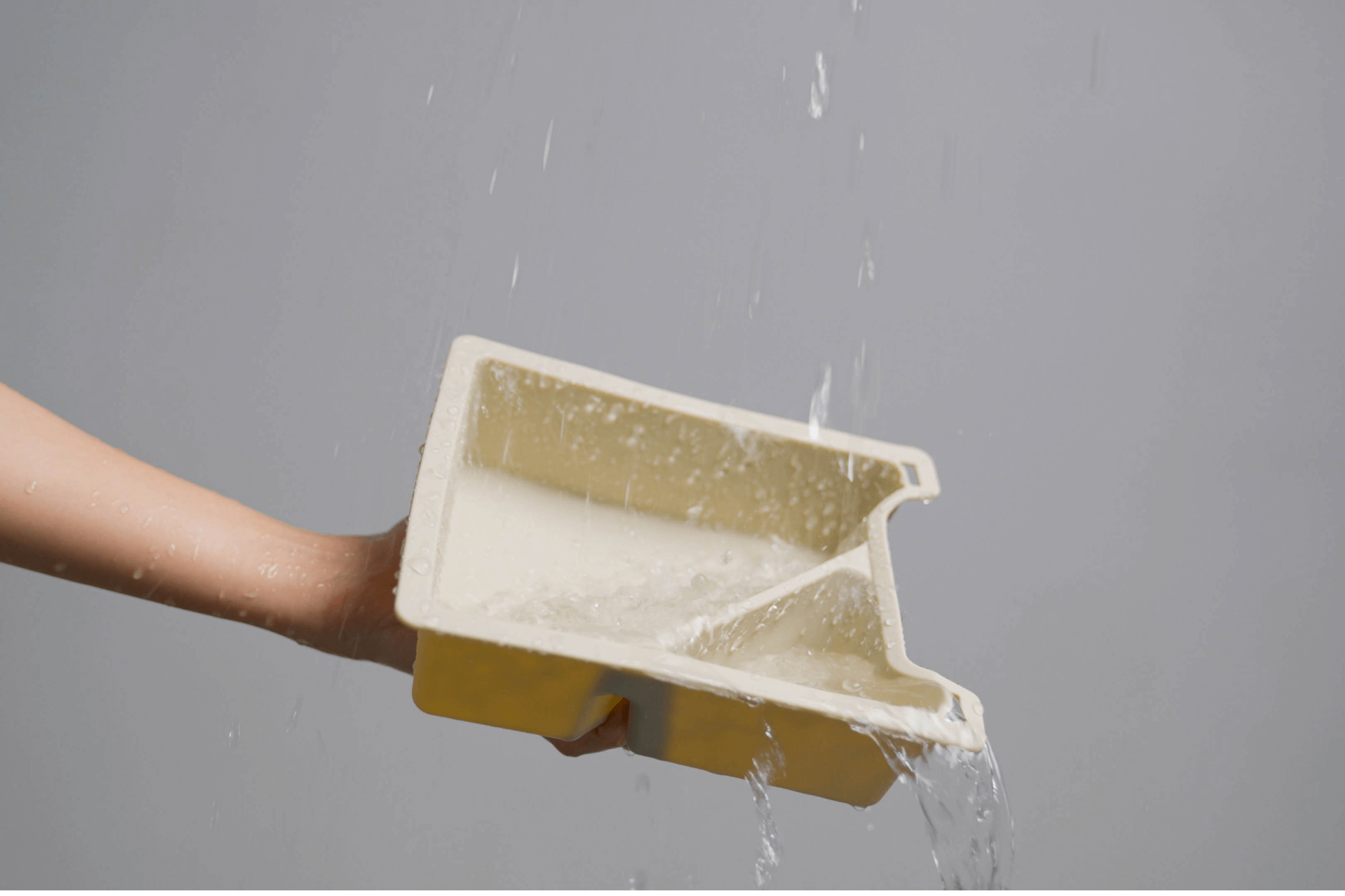 一手拿著一個黃色的長方形容器,類似 JOWUA Taiwan 中控收納盒(2021+) 的光滑雙層設計,在流水下。水從池邊濺起,背景為純灰色。.
