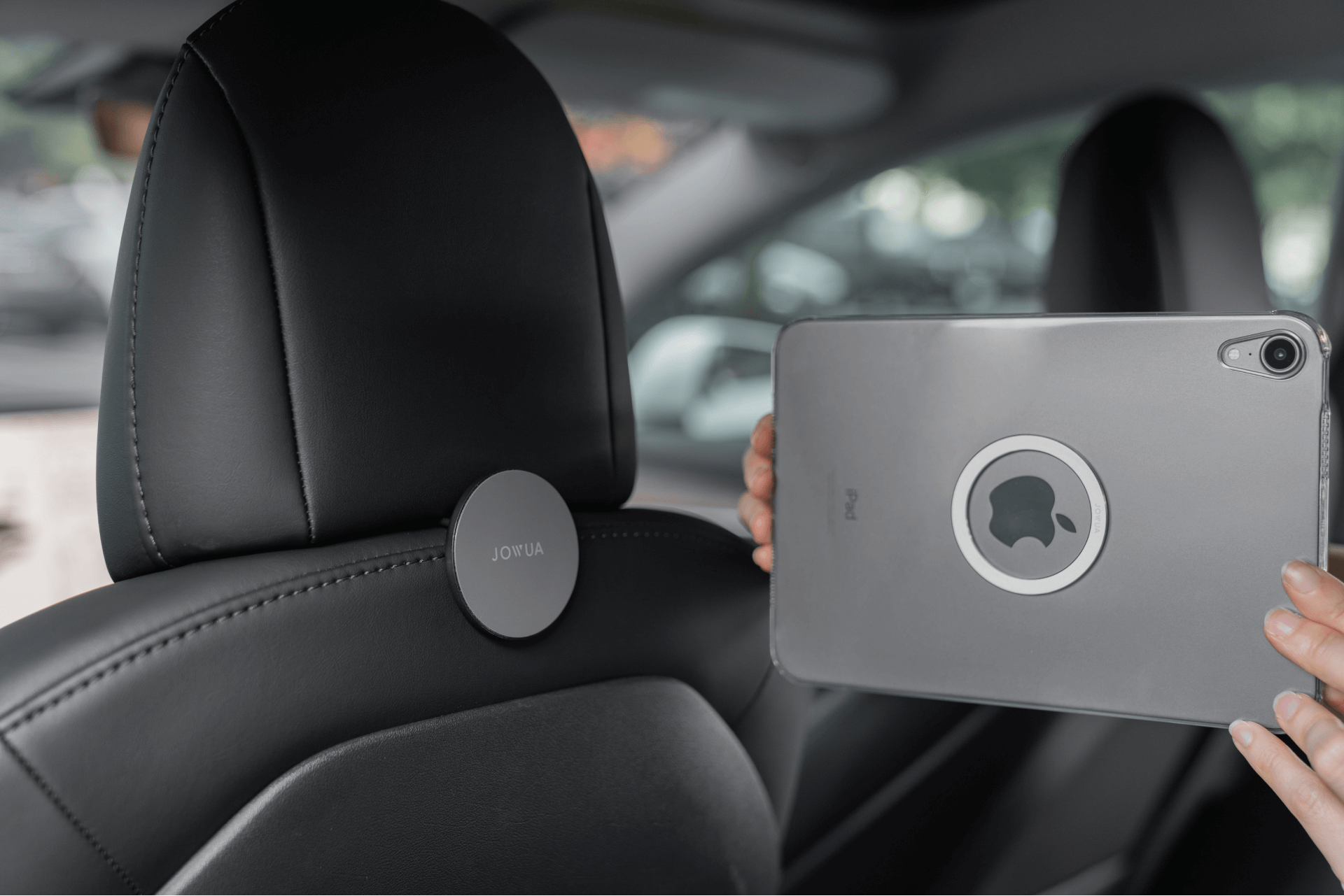 特寫一張汽車座椅的照片,其中 JOWUA Taiwan 的 MagSafe 磁吸掛鎛鉤(Model 3 2018-2024/Model Y 2021-2024)安裝在頭枕上,同時一個人拿著帶有磁吸掛鉤的 iPad,靠近掛鉤,最大限度地擴大了車內空間。.