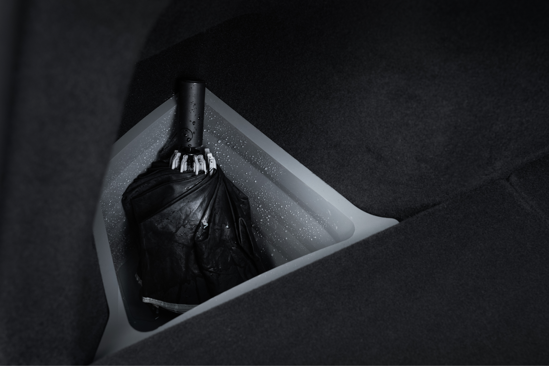 一把濕漉漉的折疊黑色雨傘放在一輛 JOWUA 台灣後行李箱收藏組(型號 Y 2021-2024)中,其三角形隔間內襯有水滴,周圍是深色織物。.