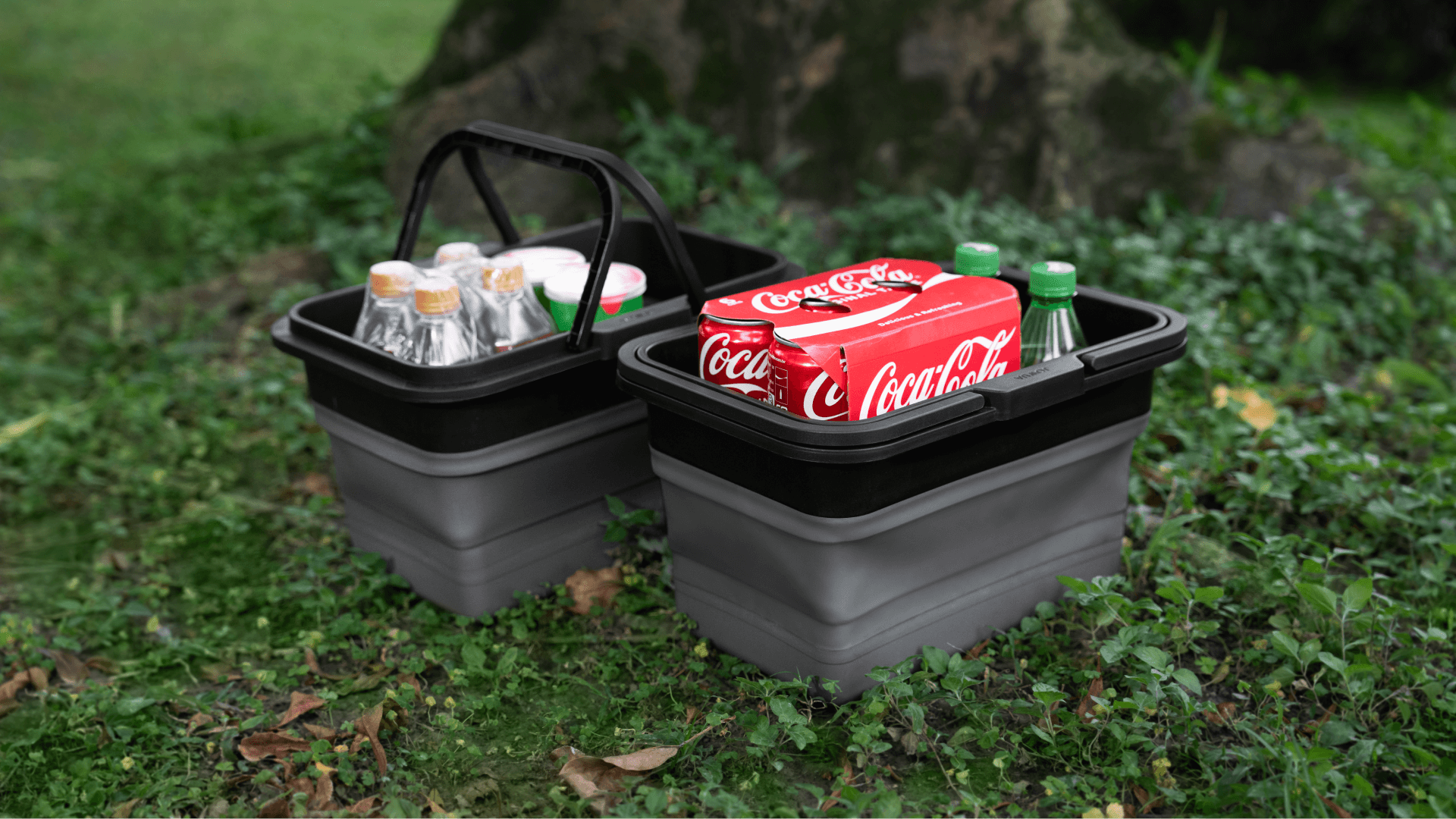 草地上擱著兩個可折疊的JOWUA台灣折疊收納盒:一個放著裝有透明液體和金色瓶蓋的瓶子,另一個放著六罐可口可樂罐和兩個綠色玻璃瓶。它們非常適合 Tesla Model X/Y,在茂密的樹叢中利用座椅下的儲存空間。.