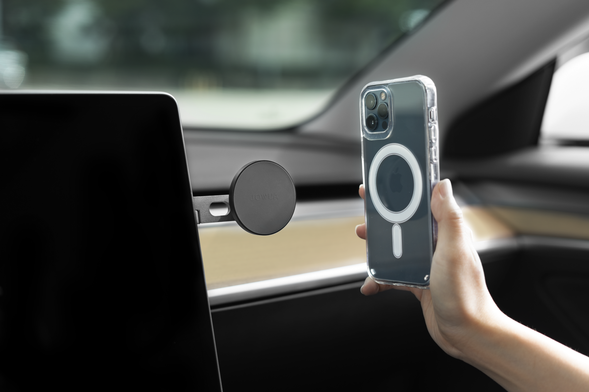一個人拿著帶有圓形磁性附件的 JOWUA 台灣 MagSafe 透明保護殼(適用於 iPhone 12/11),靠近汽車磁力支架。內裝採用木質鑲板和儀表板螢幕,具有優雅和實用的功能以及無線網路充電功能。.