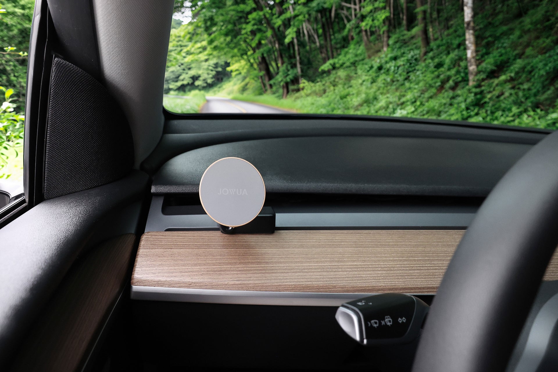 一台 JOWUA Taiwan Qi2 製冷出風口 MagSafe 車架 (Model 3/Y 2018-2024) 安裝在木質紋理的汽車儀表板上，透過車窗可以看到綠色的森林和道路。.