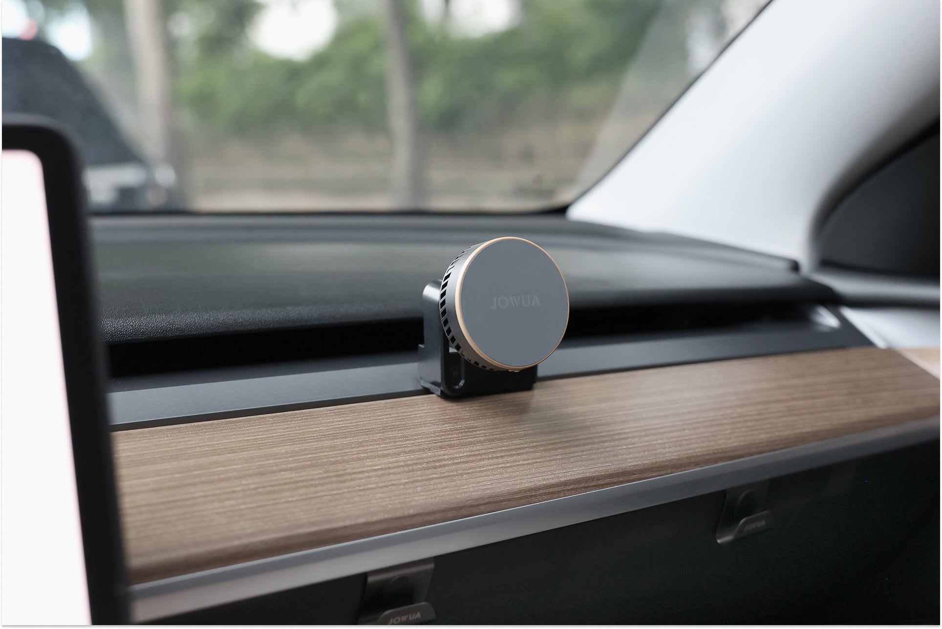 圓形、黑色和銀色的 JOWUA Taiwan Qi2 製冷出風口 MagSafe 車架 (Model 3/Y 2018-2024) 裝在汽車的木質儀表板飾板上，背景是透過擋風玻璃可見的樹木。.