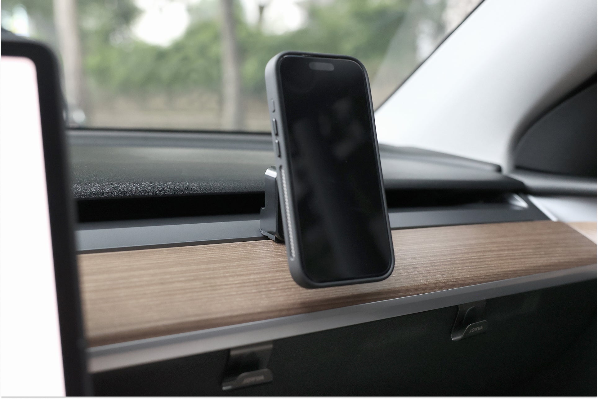黑色外殼的智慧型手機安裝在 JOWUA Taiwan Qi2 製冷出風口 MagSafe 車架 (Model 3/Y 2018-2024) 上，附在木質鑲邊的汽車儀表板上，透過擋風玻璃可以看見模糊的樹木。.