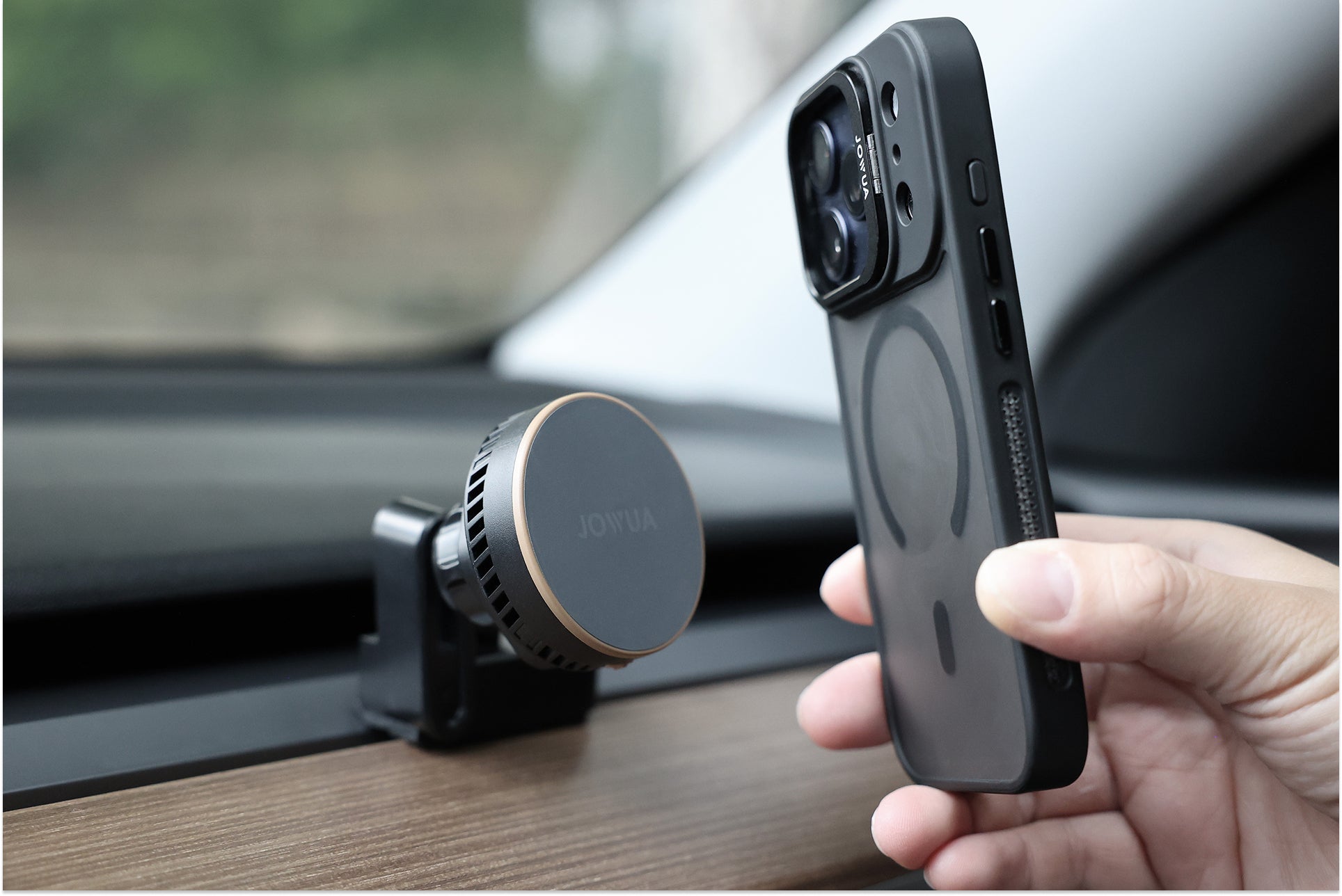 一隻手拿著一個 MagSafe 環附近的黑色 JOWUA Taiwan Qi2 製冷出風口 MagSafe 車架 (Model 3/Y 2018-2024)，穩固地固定在木飾的汽車儀表板上。.