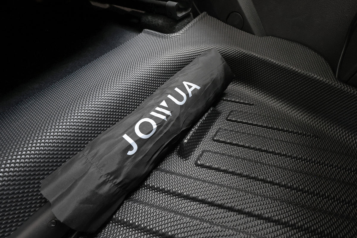 車內放置著一把印有「JOINUA」字樣的黑色雨傘，倚靠在 JOWUA 台灣出品的立體防水腳踏墊（Foxtron Cavira）上，為您的車內空間增添了風格與實用性。.