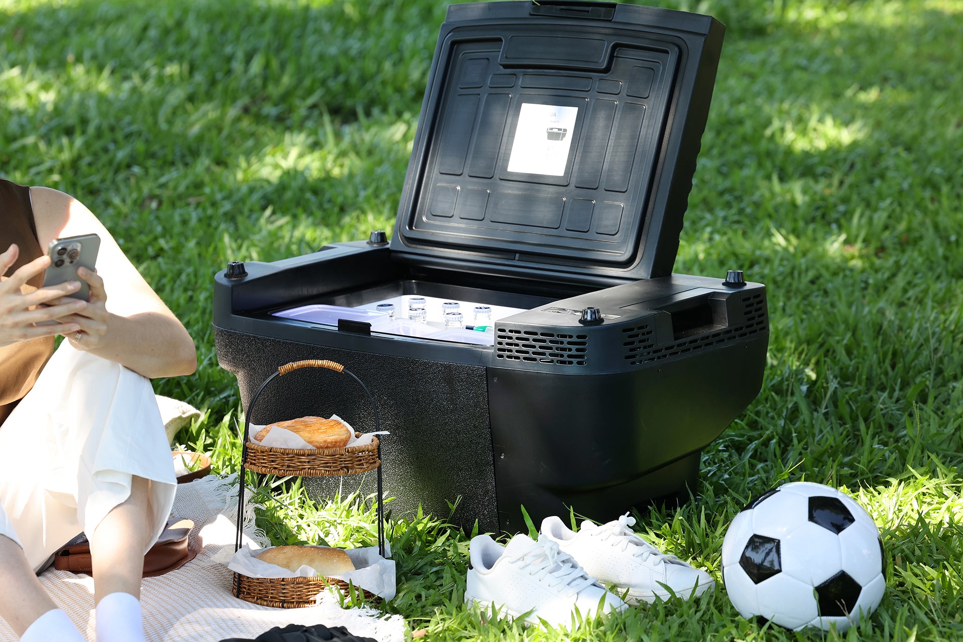 以下是根據您的產品資料重新撰寫的句子： 一款黑色款的 JOWUA 台灣製 Type-C ＆ 點菸器 雙軌車用行動冰箱（Model Y 全車系），憑藉其雙電源系統設計，具備多功能用途，此刻正敞開著置於草地上，旁邊放著一個足球、一雙白色運動鞋、一個裝滿麵包和糕點的野餐籃，以及一位正在使用智慧型手機的人。.