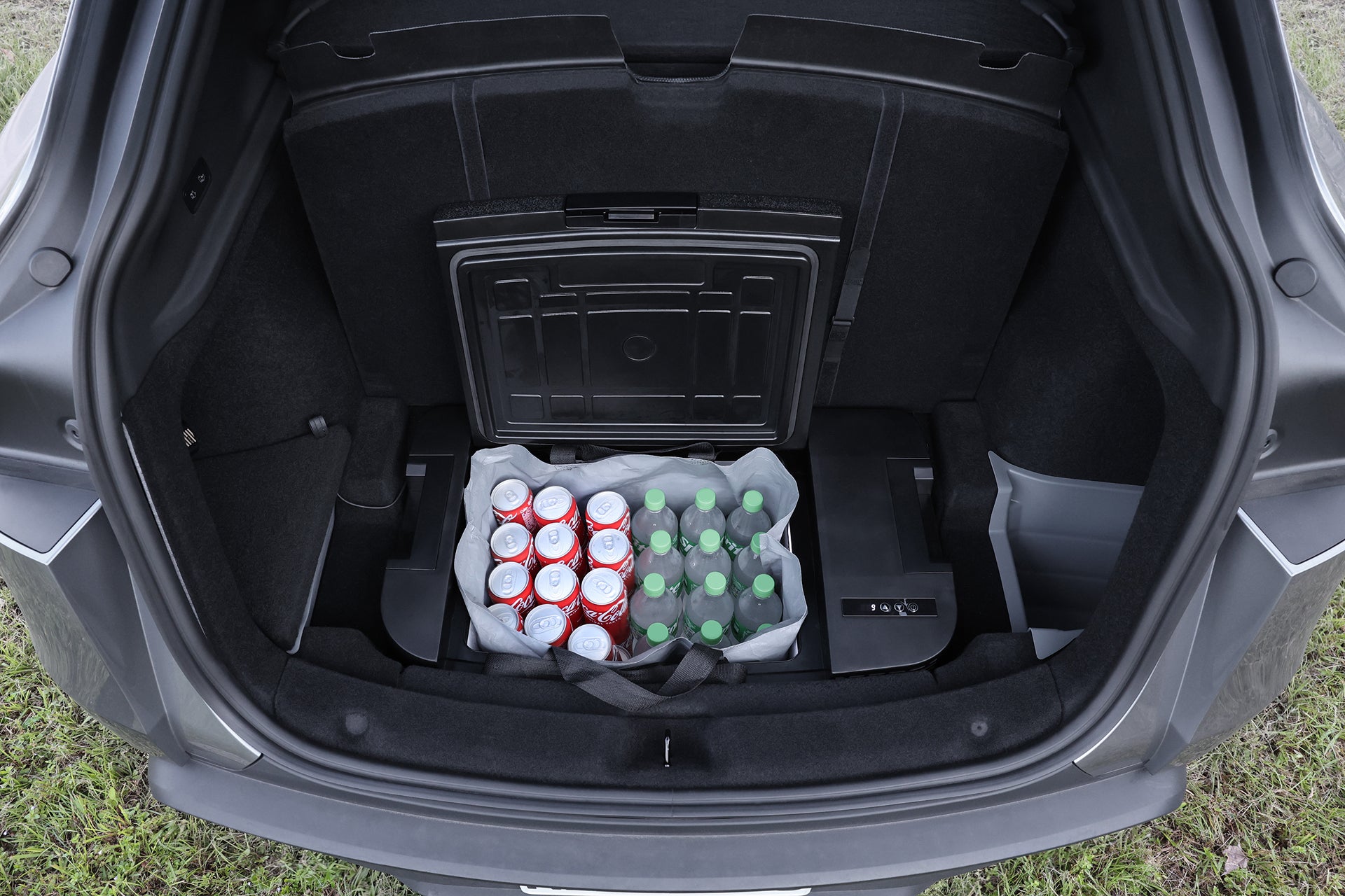 打開後車廂可看到 JOWUA Taiwan Type-C＆點菸器雙軌車用行動冰箱（Model Y全車系）35L大容量，後車廂地板上有一個可重複使用的袋子，裡面裝有汽水罐和水樽，外面可見草地。.