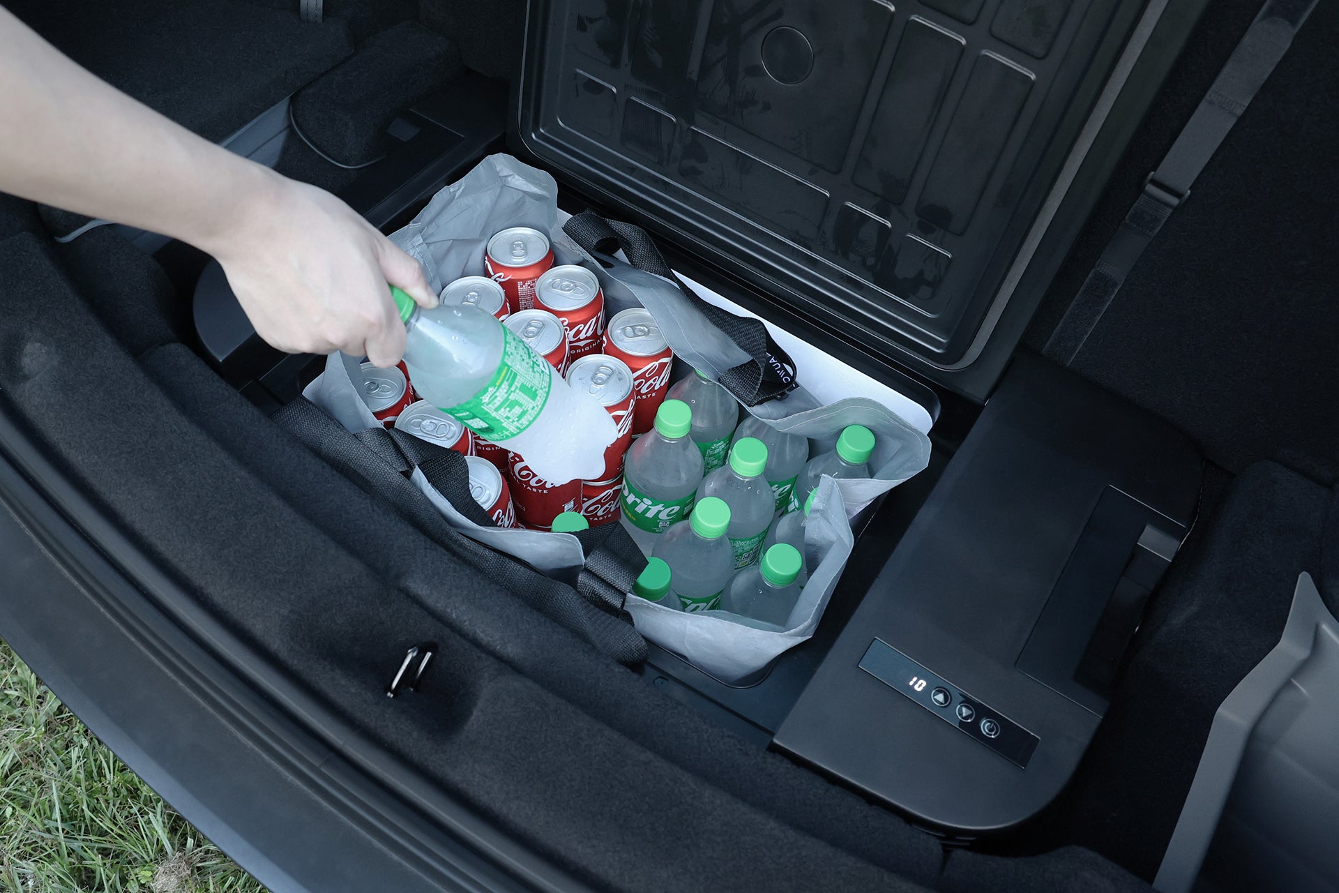 一隻手將綠色瓶蓋的汽水瓶放入灰色的 JOWUA Taiwan Type-C & 點菸器雙軌車用行動冰箱 (Model Y全車系)，後車廂則裝滿飲料與可口可樂罐，確保旅途中的清涼飲料。.