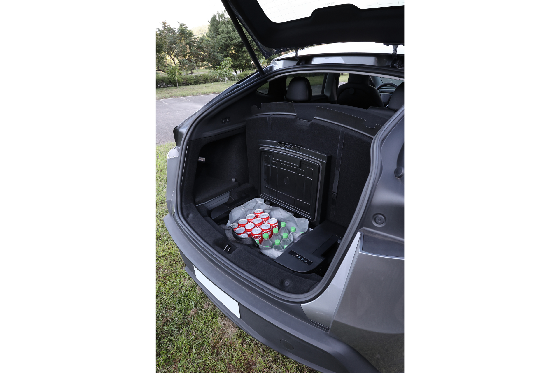 打開的休旅車後備箱中，有一台 35L 的 JOWUA Taiwan Type-C & 點菸器 雙軌車用行動冰箱 (Model Y全車系)，裡頭擺滿了飲料，停在路旁的草地上，背景是綠意盎然的樹木。.