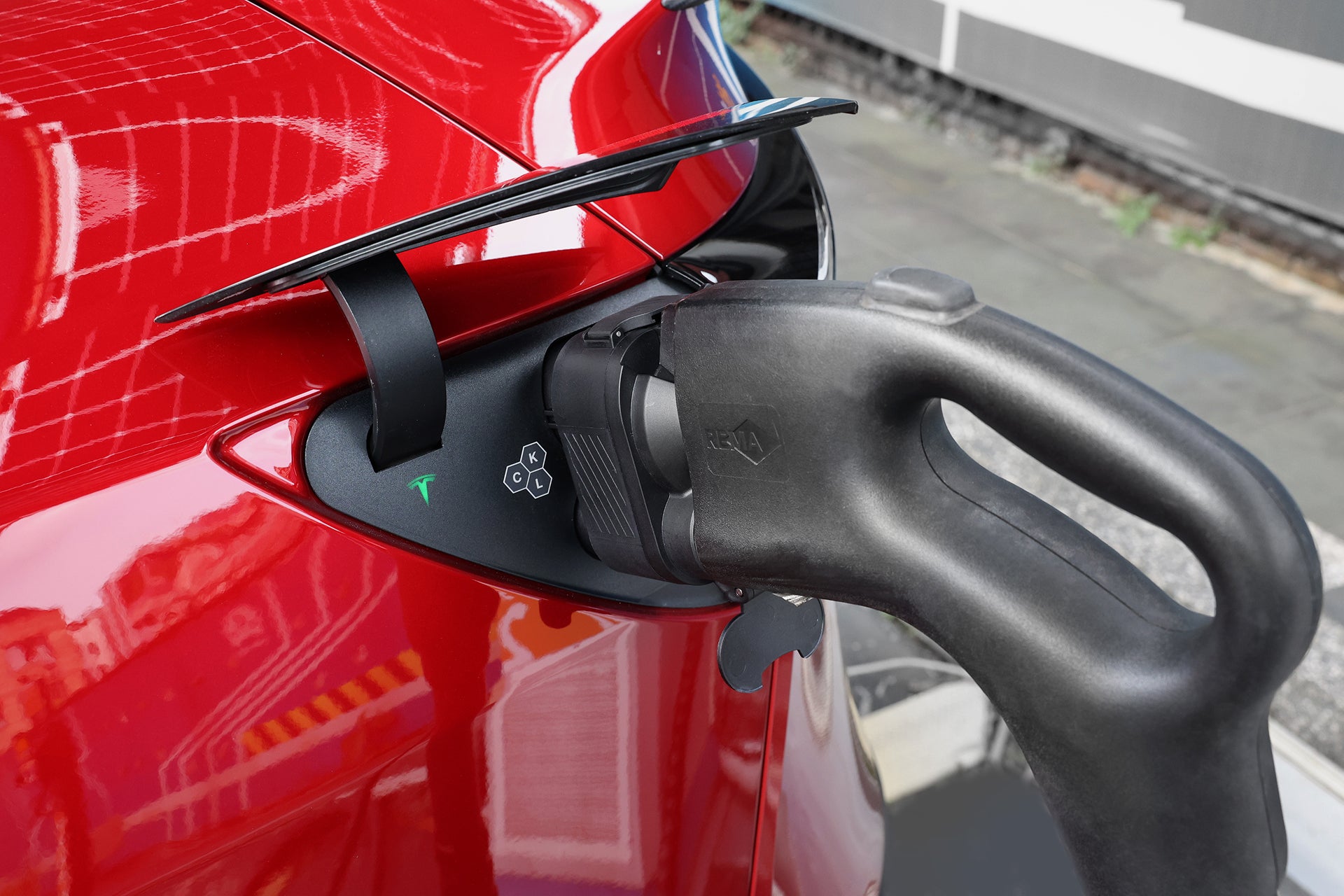 一張閃亮的紅色電動車特寫照片，該車正透過 JOWUA 台灣製造的 CCS1 轉 CCS2 直流與交流快慢充轉接頭，以黑色插頭進行充電。背景中可見路面及建築物的一部分。.